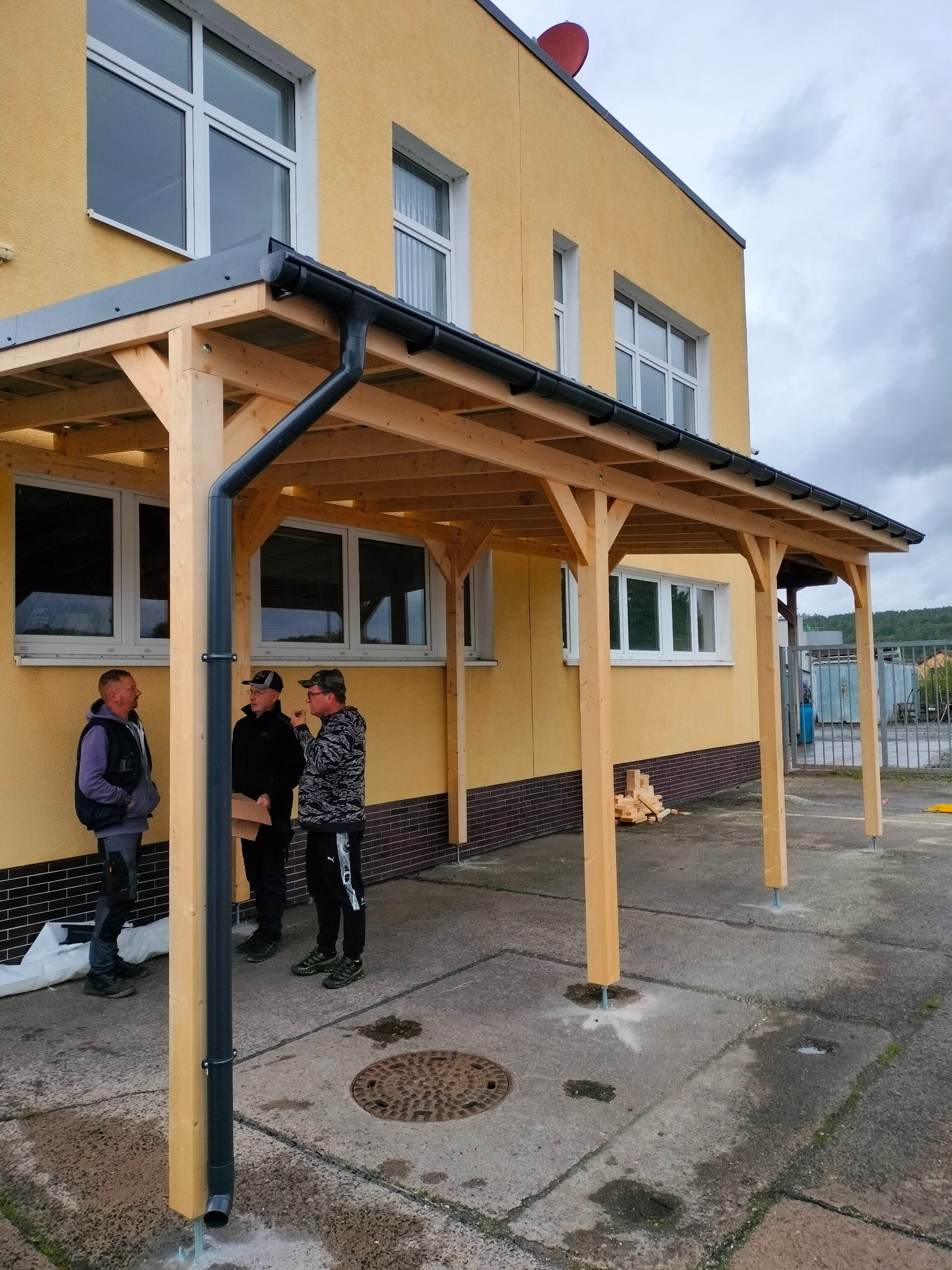 Drewniana wiata z rynną przy żółtym budynku. Trzy osoby rozmawiają obok konstrukcji. Widoczne elementy montażowe i stos drewna.