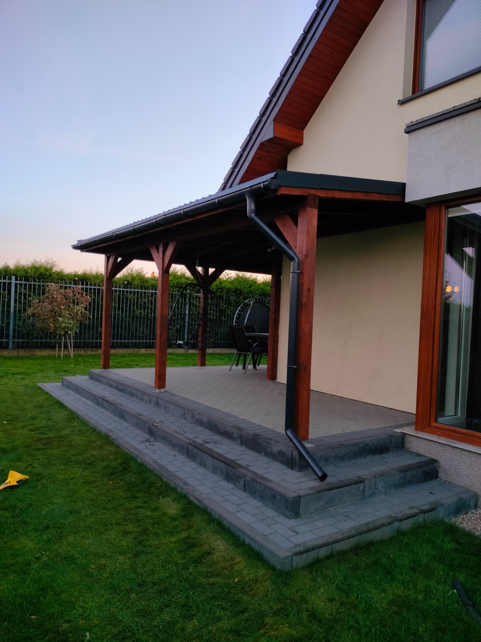 Drewniana, zadaszona konstrukcja tarasowa przylegająca do budynku. Widoczne schody z kostki brukowej, fragment trawnika i ogrodzenie. Elementy drewniane w kolorze brązowym.