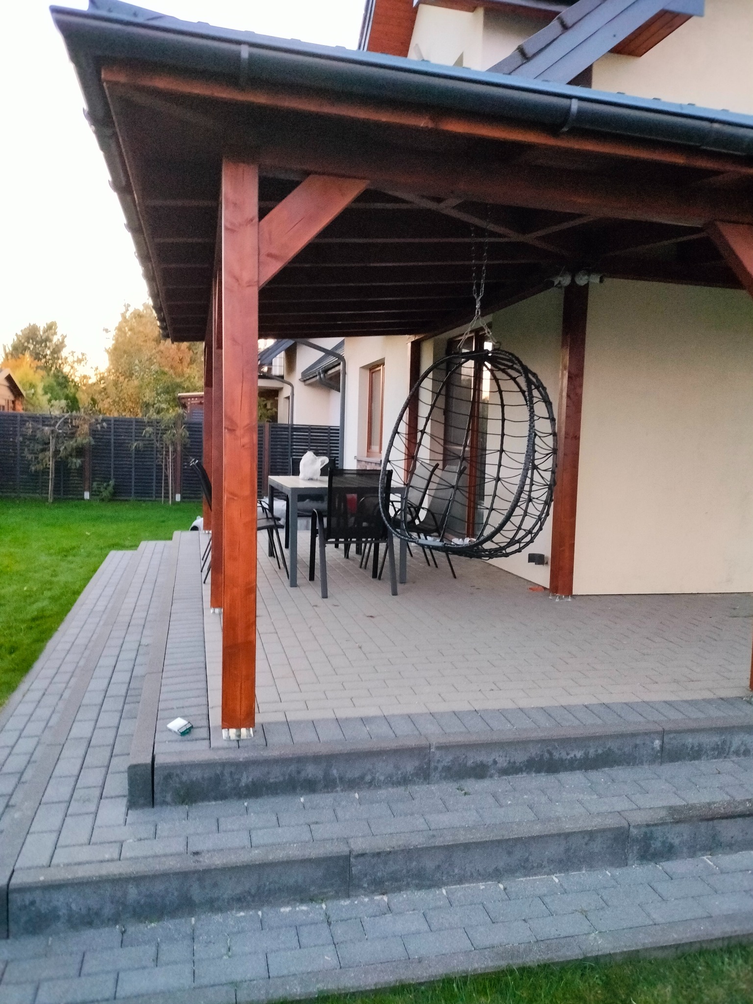 Zadaszona, kamienna weranda z drewnianymi słupami. W tle dom z oknem. Na werandzie stół z krzesłami i wiszący fotel. Widoczne schody z kostki brukowej i trawnik.