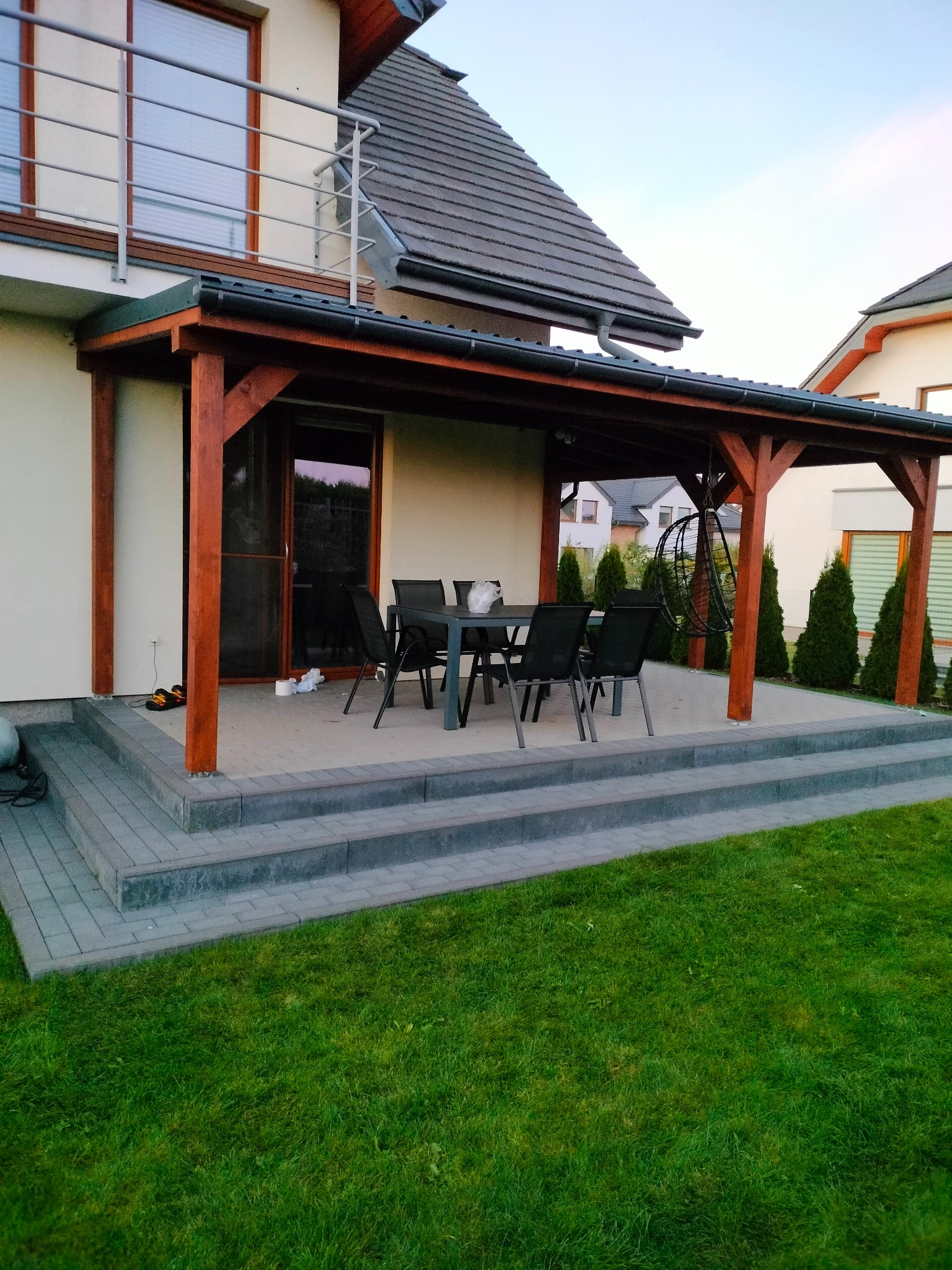 Zadaszony taras z drewnianą konstrukcją i schodami z kostki brukowej, wyposażony w stół i krzesła. W tle zielony trawnik i tuje.