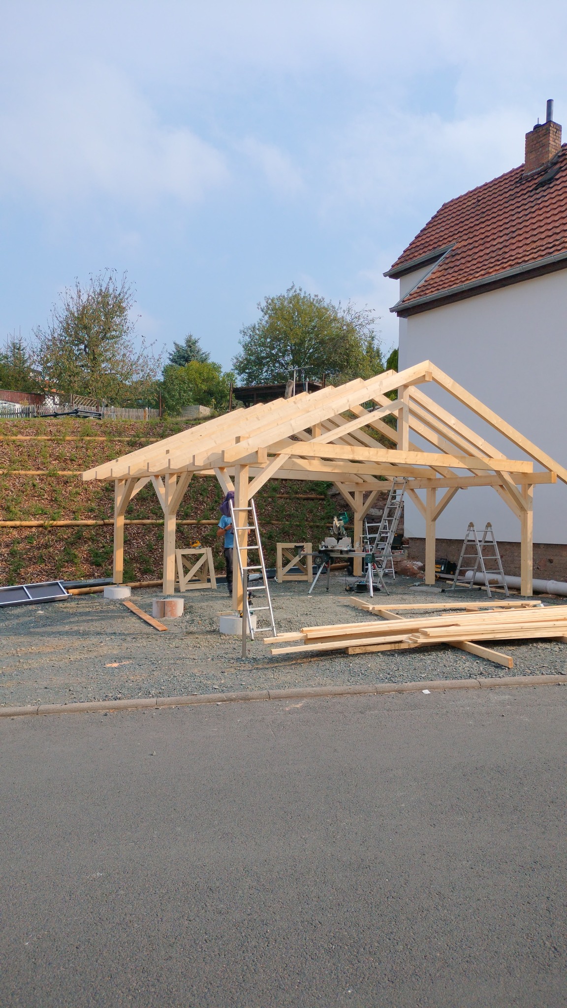 Konstrukcja drewniana z jasnego drewna w trakcie budowy, widoczne narzędzia i drabiny, dom w tle, słoneczny dzień.