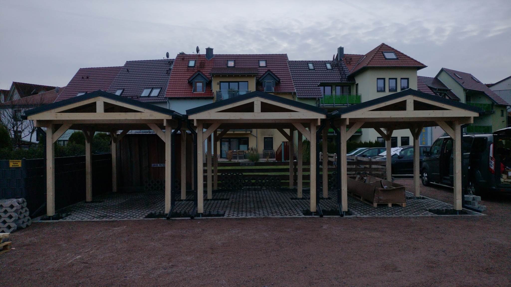 Trzy drewniane, połączone wiaty garażowe z czarnymi dachami na posesji z kostką ażurową, na tle budynków mieszkalnych w pochmurny dzień.