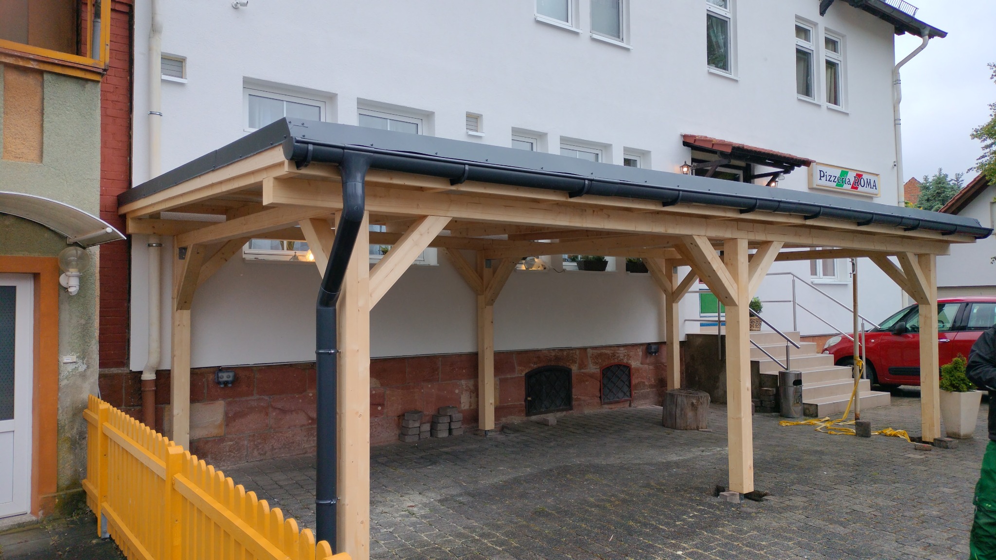 Nowoczesna, drewniana konstrukcja z szarą blachą trapezową, prawdopodobnie zadaszenie lub wiata, przylegająca do budynku z oknami. Widoczny system rynnowy.