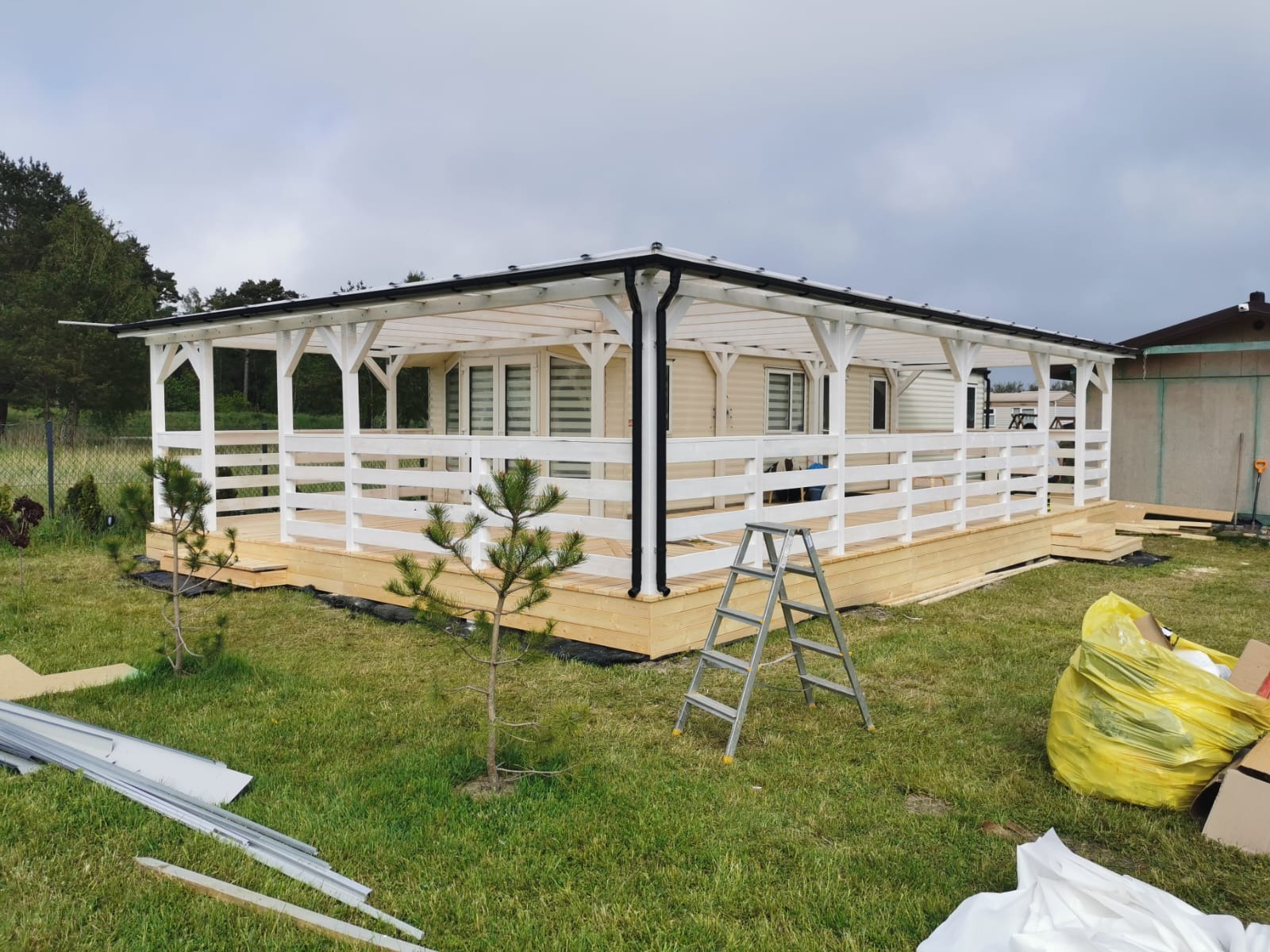 Biała altana z drewnianą podłogą na trawie, drabina oparta o konstrukcję. Widoczne elementy montażowe i torba z odpadami. Pochmurne niebo w tle.