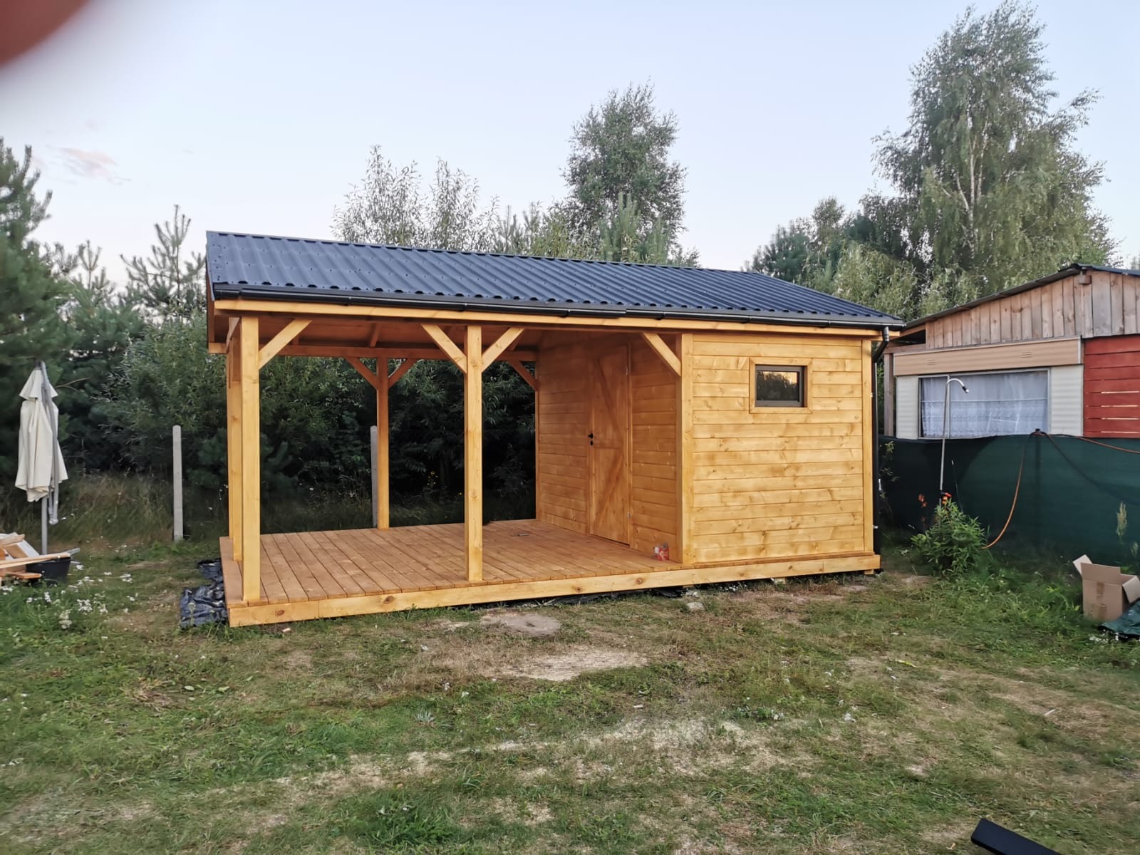 Drewniana altana z zadaszeniem z blachy trapezowej, widok z zewnątrz na trawniku. Elementy konstrukcyjne z surowego drewna, widoczne łączenia. Tło: zieleń drzew.