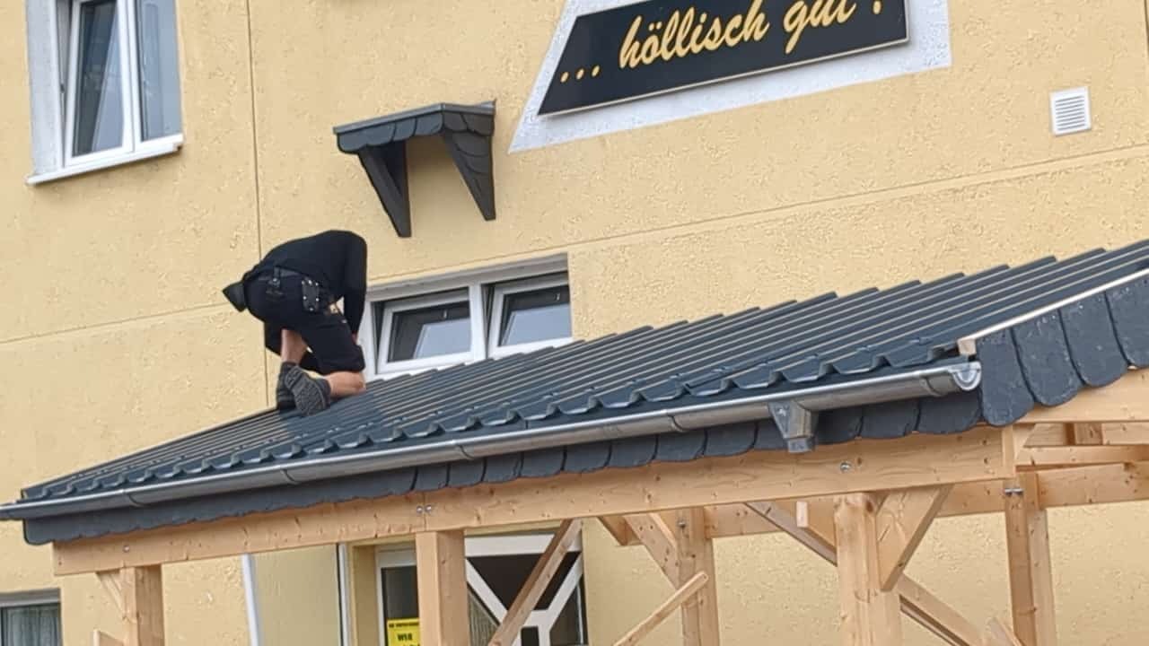 Dekarz montuje dach z blachy trapezowej na drewnianej konstrukcji. Widoczne narzędzia dekarskie i fragment budynku z oknami. Ujęcie z dołu, z bliska.