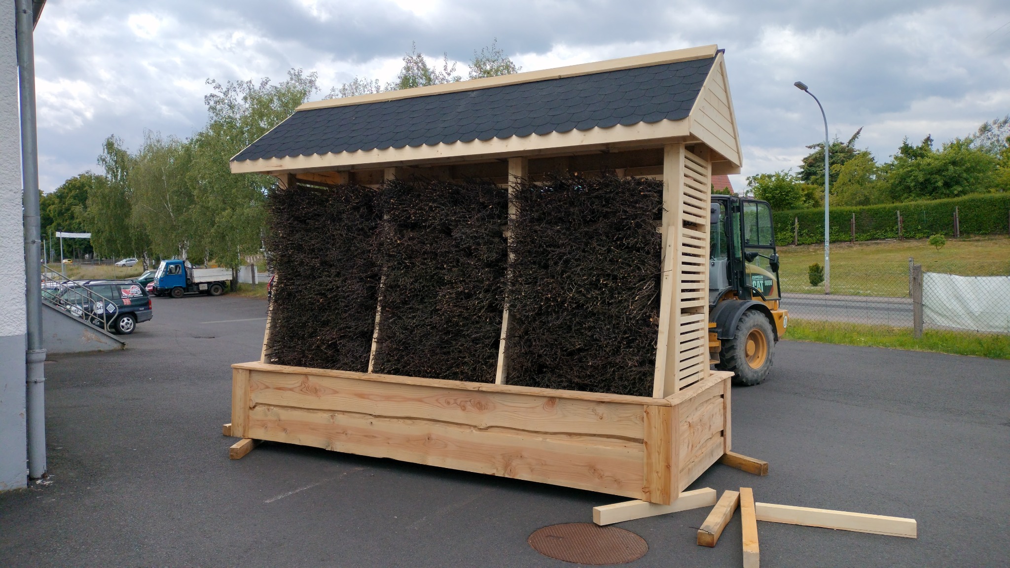 Drewniana konstrukcja z dachem krytym gontem, wypełniona gałęziami, przypominająca małą tężnię solankową, ustawiona na zewnątrz. W tle widoczny traktor.