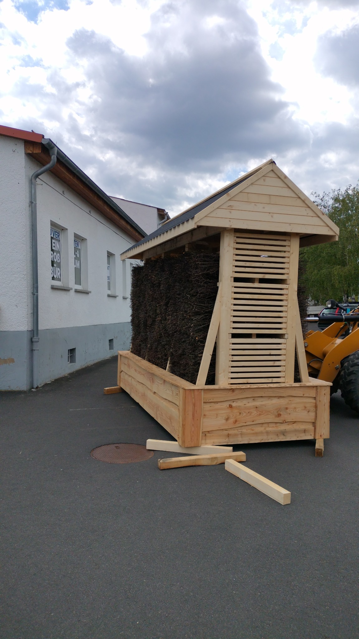 Tężnia solankowa w trakcie budowy: drewniana konstrukcja z wypełnieniem z gałęzi, widoczny budynek w tle, częściowo widoczna maszyna budowlana.