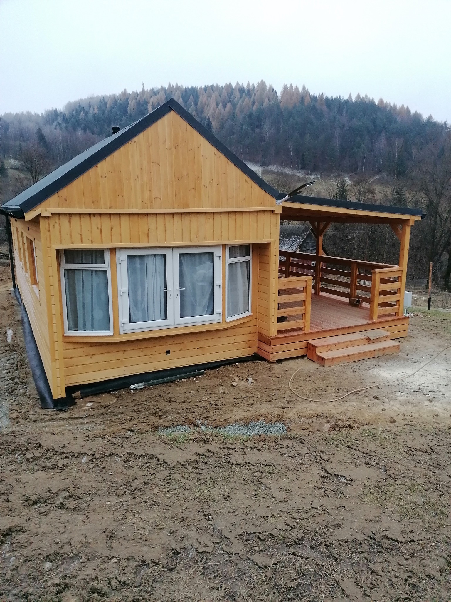 Żółty, drewniany domek z gankiem i czarnym dachem na tle zalesionych wzgórz w pochmurny dzień. Widoczne okna z zasłonami i drewniane balustrady ganku.