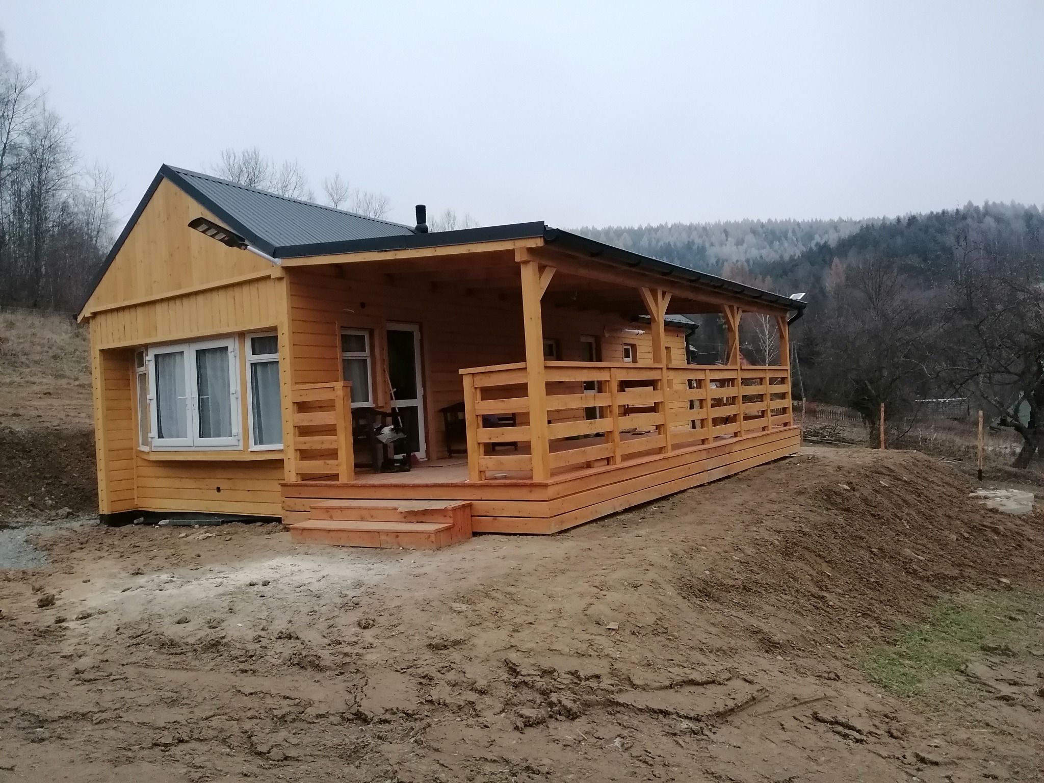 Żółty dom szkieletowy z zadaszonym tarasem i blachą trapezową na dachu, na tle zalesionych wzgórz w pochmurny dzień. Widok z boku na niezagospodarowany teren.