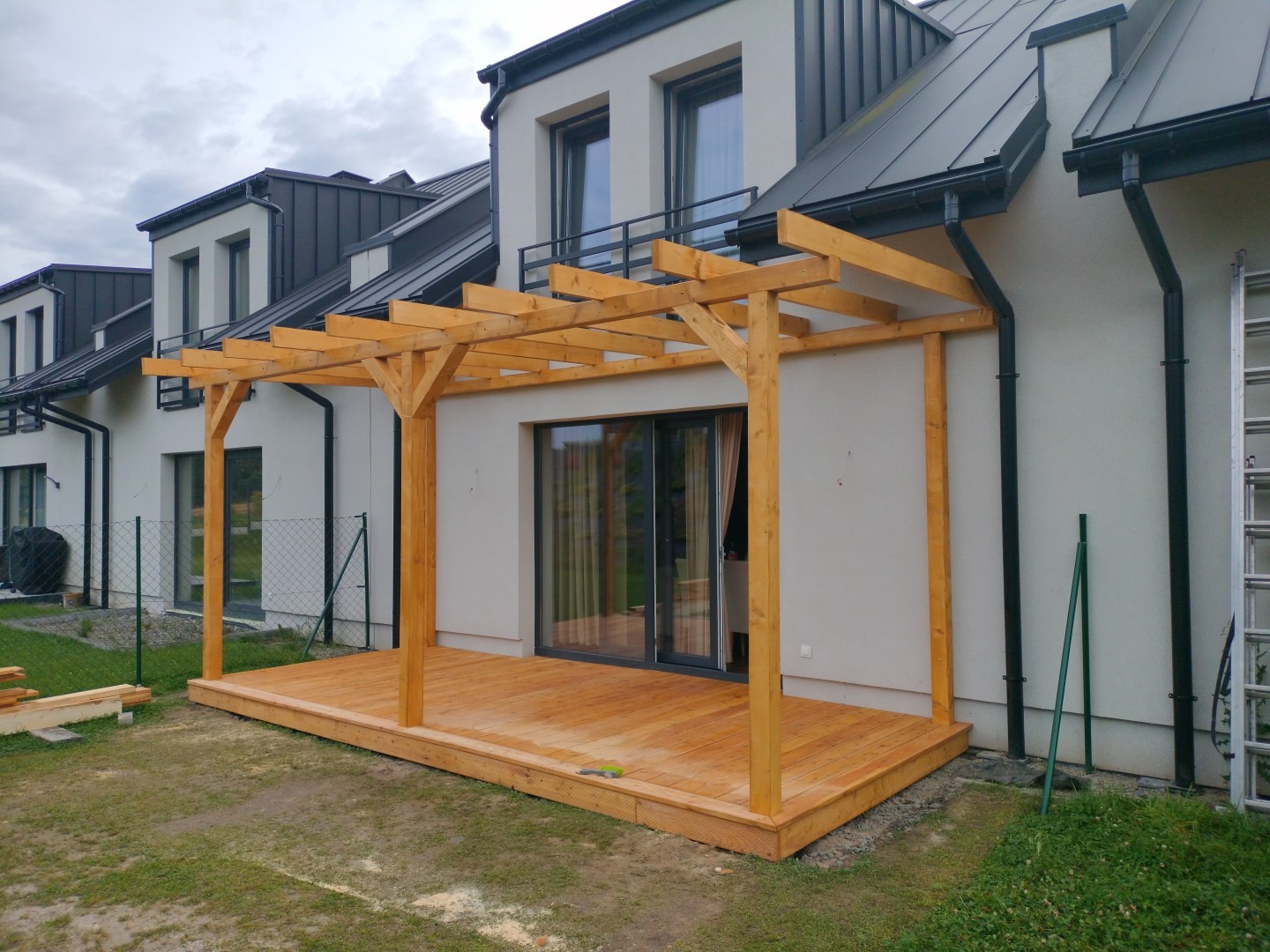 Drewniana pergola z tarasem przy nowoczesnym domu z grafitowym dachem. Jasne drewno konstrukcji kontrastuje z bielą elewacji. Widoczne złączenia ciesielskie.