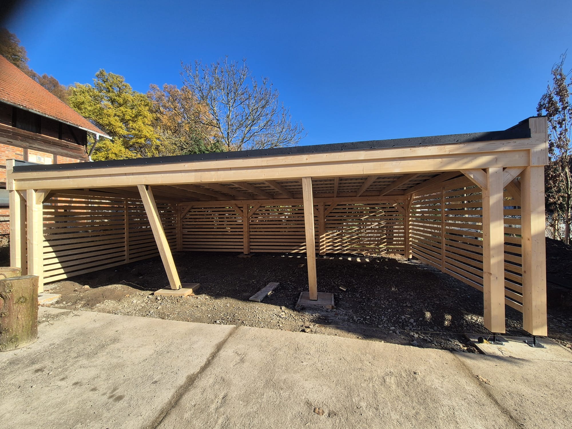 Drewniana konstrukcja z poziomymi listwami, prawdopodobnie wiata lub zadaszenie, w trakcie budowy. Widoczne elementy konstrukcyjne i podłoże z kamieni.