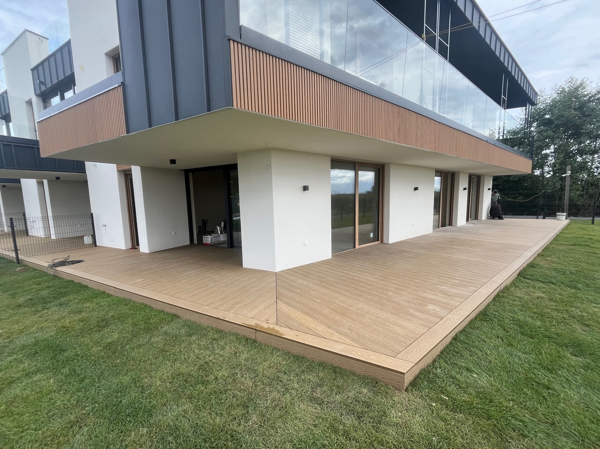 Przestronny taras z desek w odcieniach brązu, przylegający do nowoczesnego domu z dużymi oknami i elementami drewnianymi. Widok na zielony trawnik.