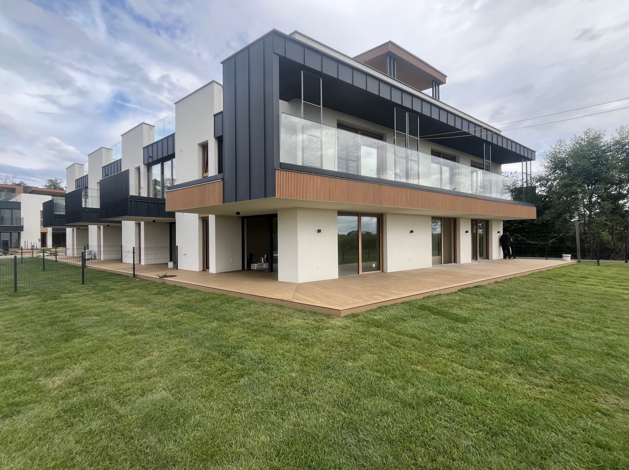 Nowoczesny, trzypiętrowy budynek z dużym tarasem na parterze, wykończonym deskami w odcieniach drewna. Widok z poziomu trawnika w słoneczny dzień.