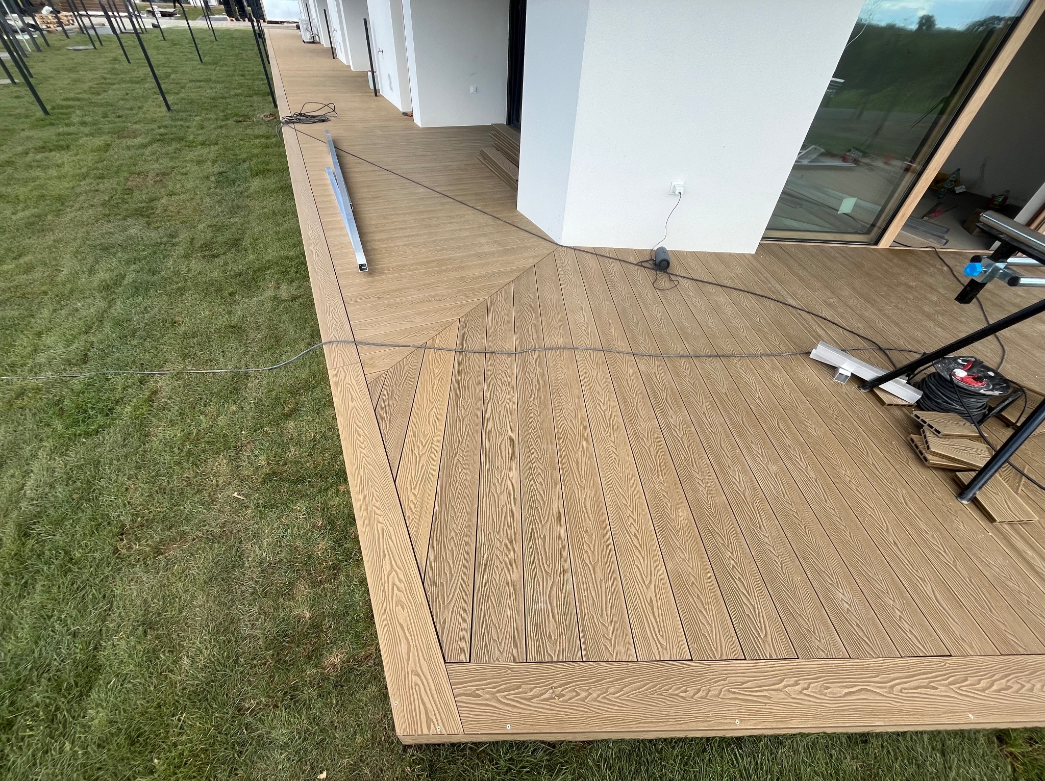 Nowoczesny taras z desek w jodełkę, częściowo ukończony, z widocznym fragmentem trawnika i narzędziami montażowymi. Jasne, naturalne oświetlenie.