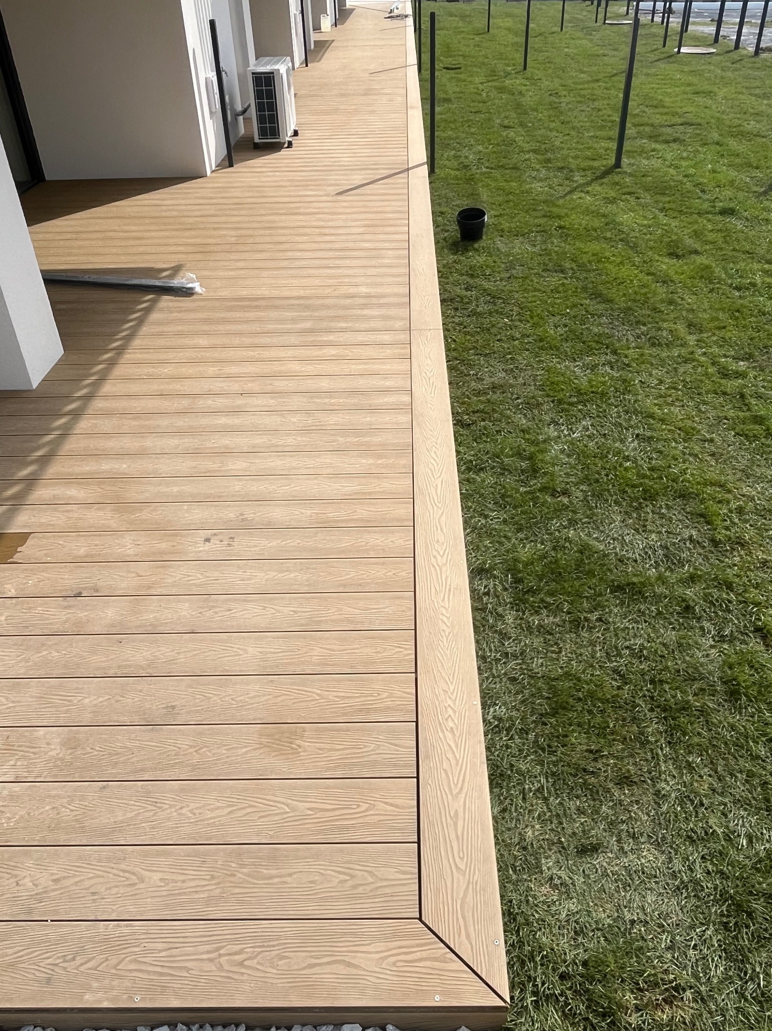 Jasny, drewnopodobny taras przy budynku z klimatyzatorem, sąsiadujący z zielonym trawnikiem i czarnymi słupkami. Widok z góry.