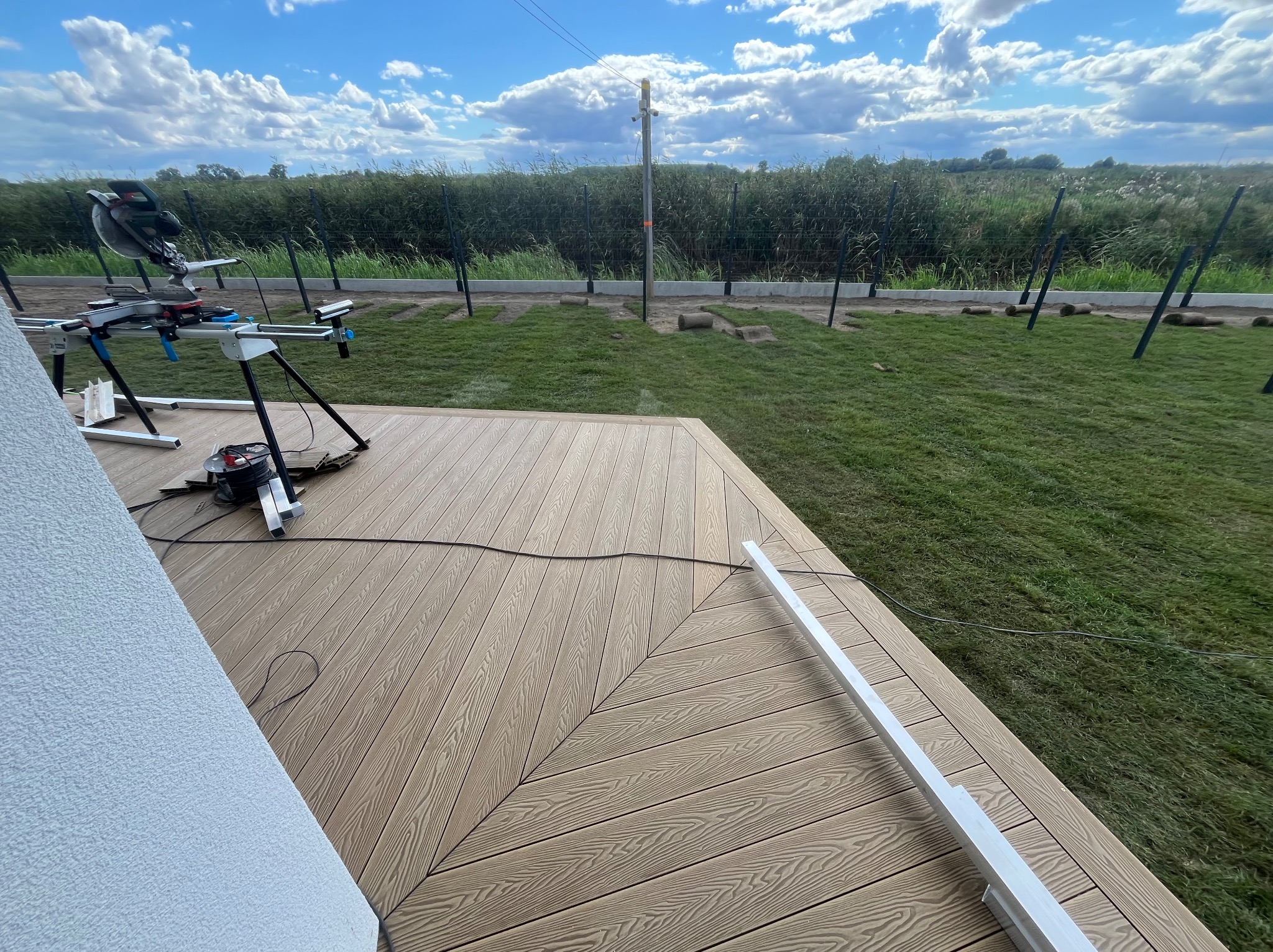 Montaż tarasu kompozytowego: deski ułożone w jodełkę, widoczna piła do cięcia, trawa w rolkach przygotowana do ułożenia. Etap prac.