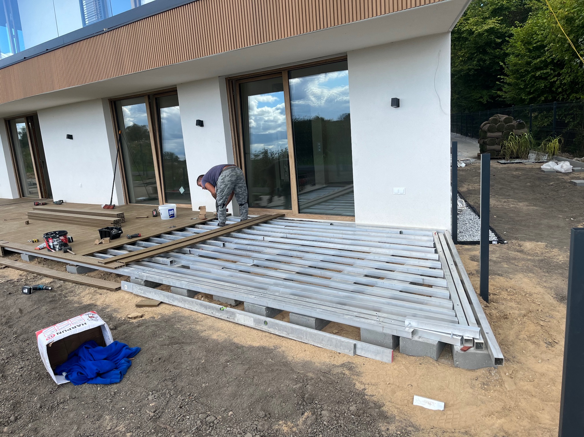 Montaż stelaża pod podłogę tarasu przy nowoczesnym domu. Widoczne legary, poziomica, narzędzia i częściowo ułożone deski. Prace w toku, widok z boku.