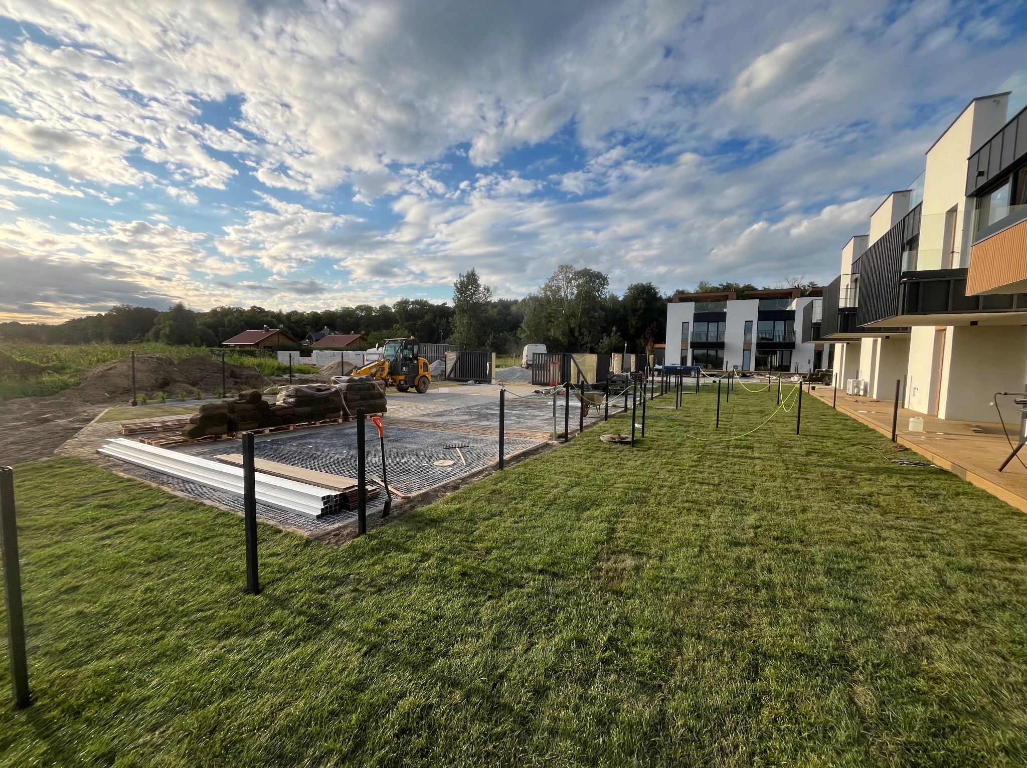 Przygotowanie podłoża pod taras: wyrównany teren, metalowa konstrukcja, rury, trawa, w tle budynki i koparka, błękitne niebo z chmurami.