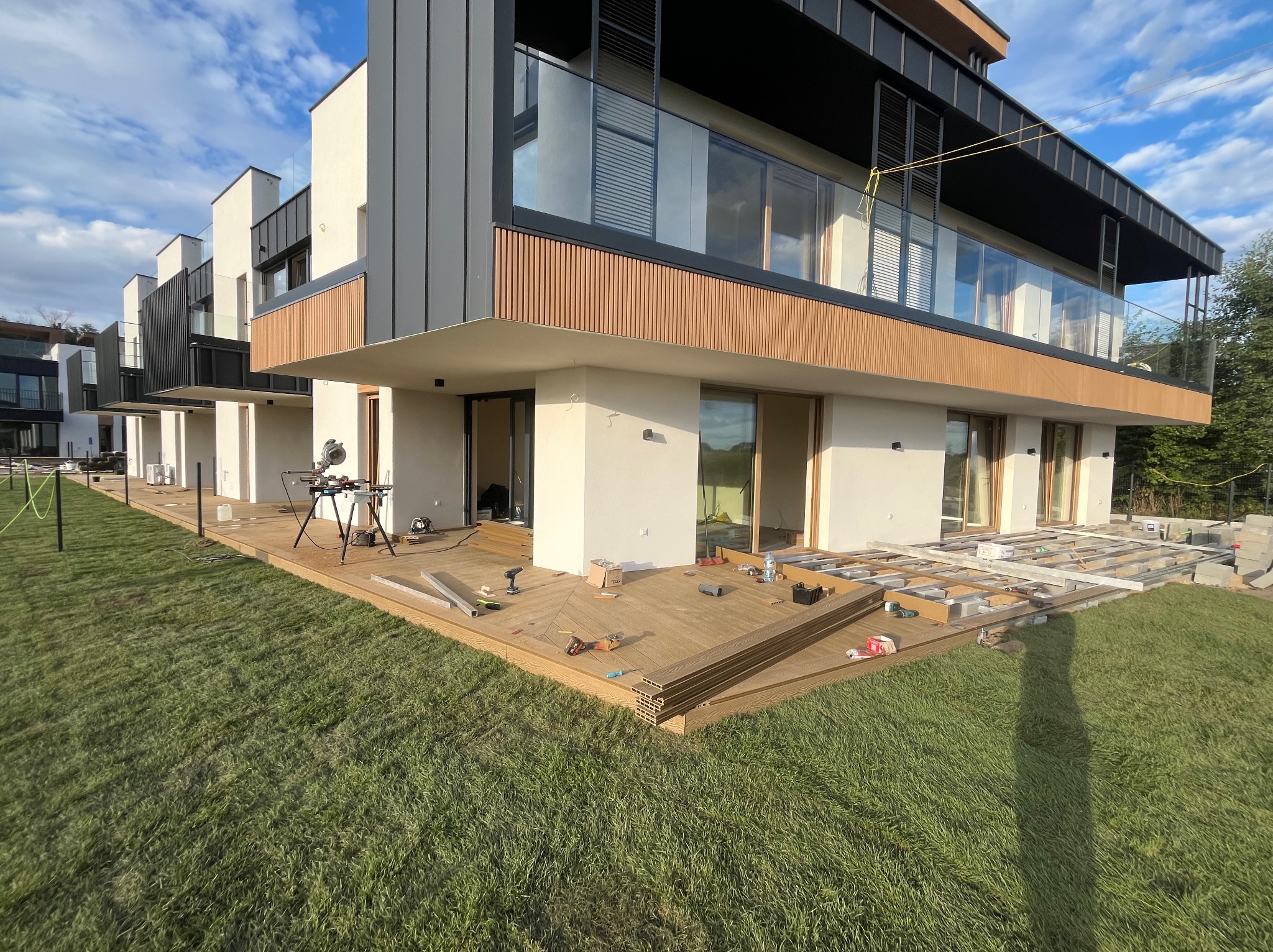 Montaż desek na tarasie przy nowoczesnym domu. Widoczne narzędzia, częściowo ułożona podłoga i trawnik. Jasne niebo w tle. Ujęcie z poziomu trawy.