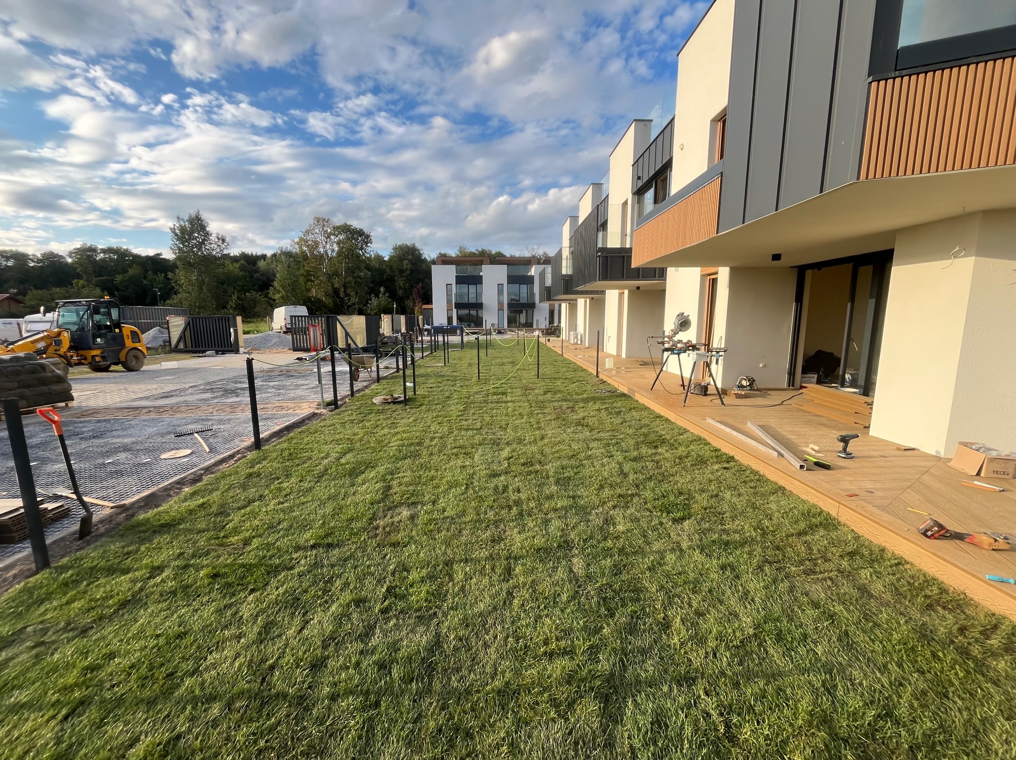 Montaż tarasu przy nowoczesnym domu z trawnikiem. Widoczne narzędzia i deski. W tle mini koparka i materiały budowlane. Słoneczny dzień.