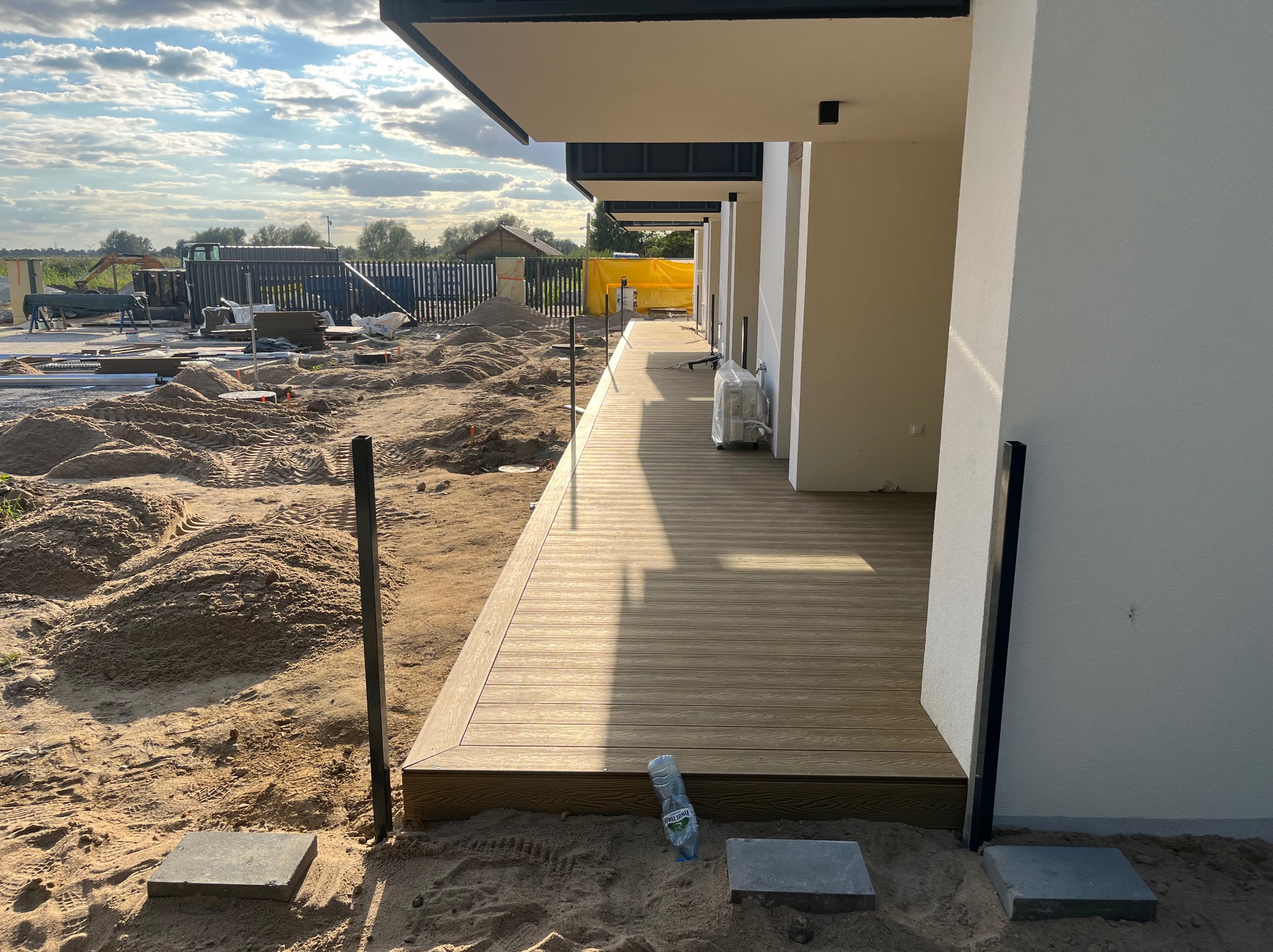 Jasna, drewniana podłoga tarasu w trakcie budowy, z widocznym piaskiem i elementami konstrukcyjnymi. W tle dom z białymi ścianami i fragment krajobrazu.