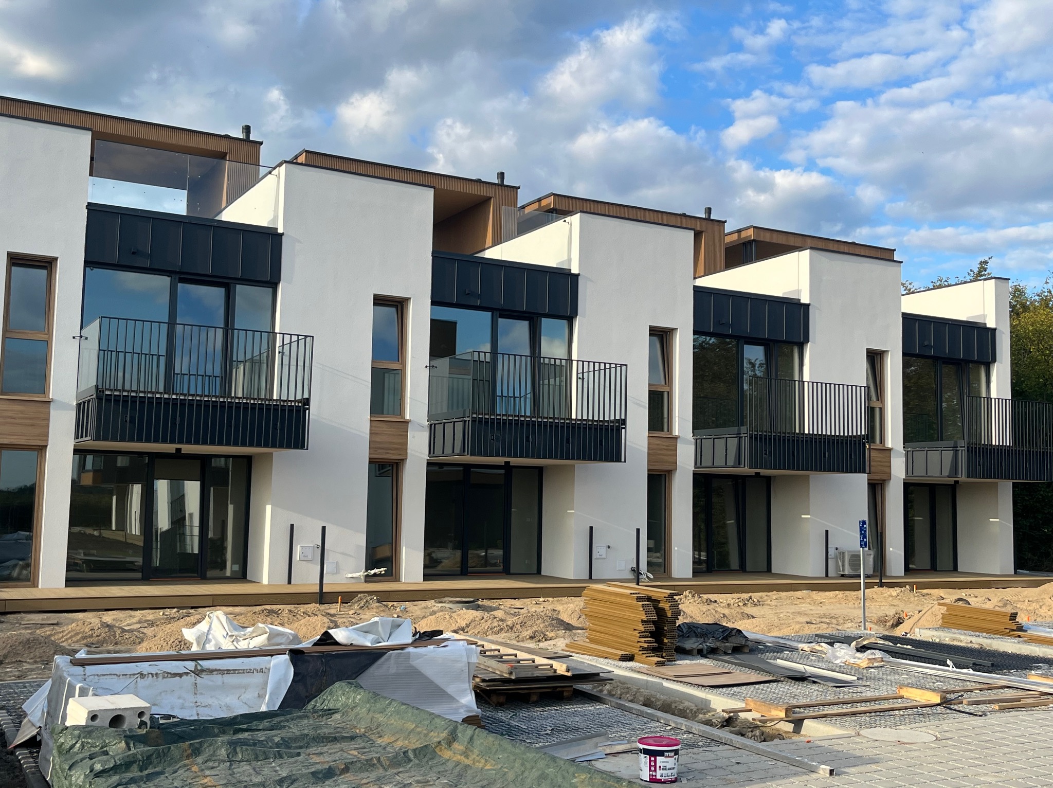 Nowoczesne, białe domy z tarasami w trakcie budowy. Widoczne deski tarasowe i materiały budowlane. Błękitne niebo z chmurami w tle.