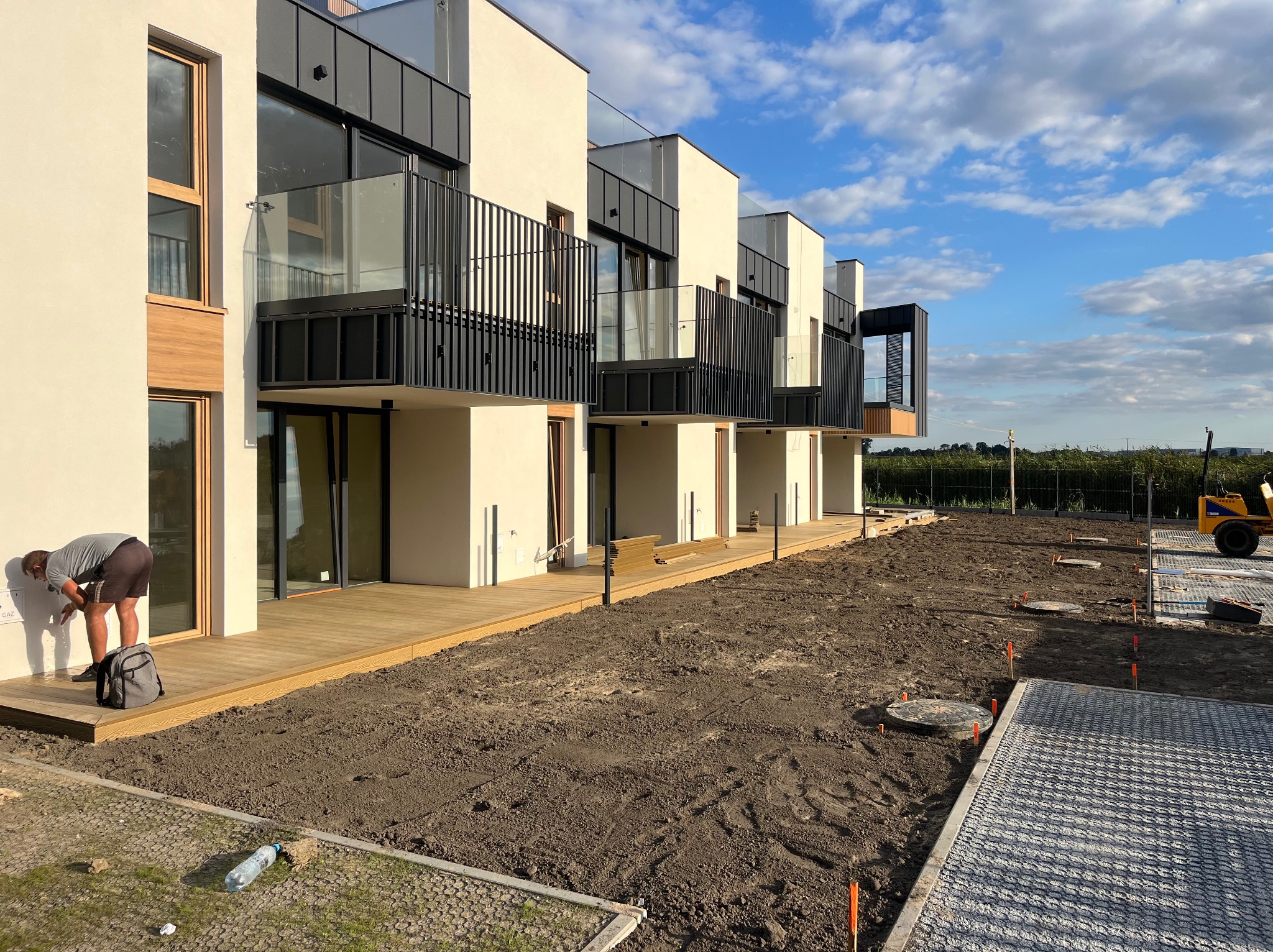 Nowoczesne tarasy przy szeregu domów w trakcie budowy. Widoczny pracownik przy elewacji, drewniane podesty i przygotowany teren pod dalsze prace wykończeniowe.