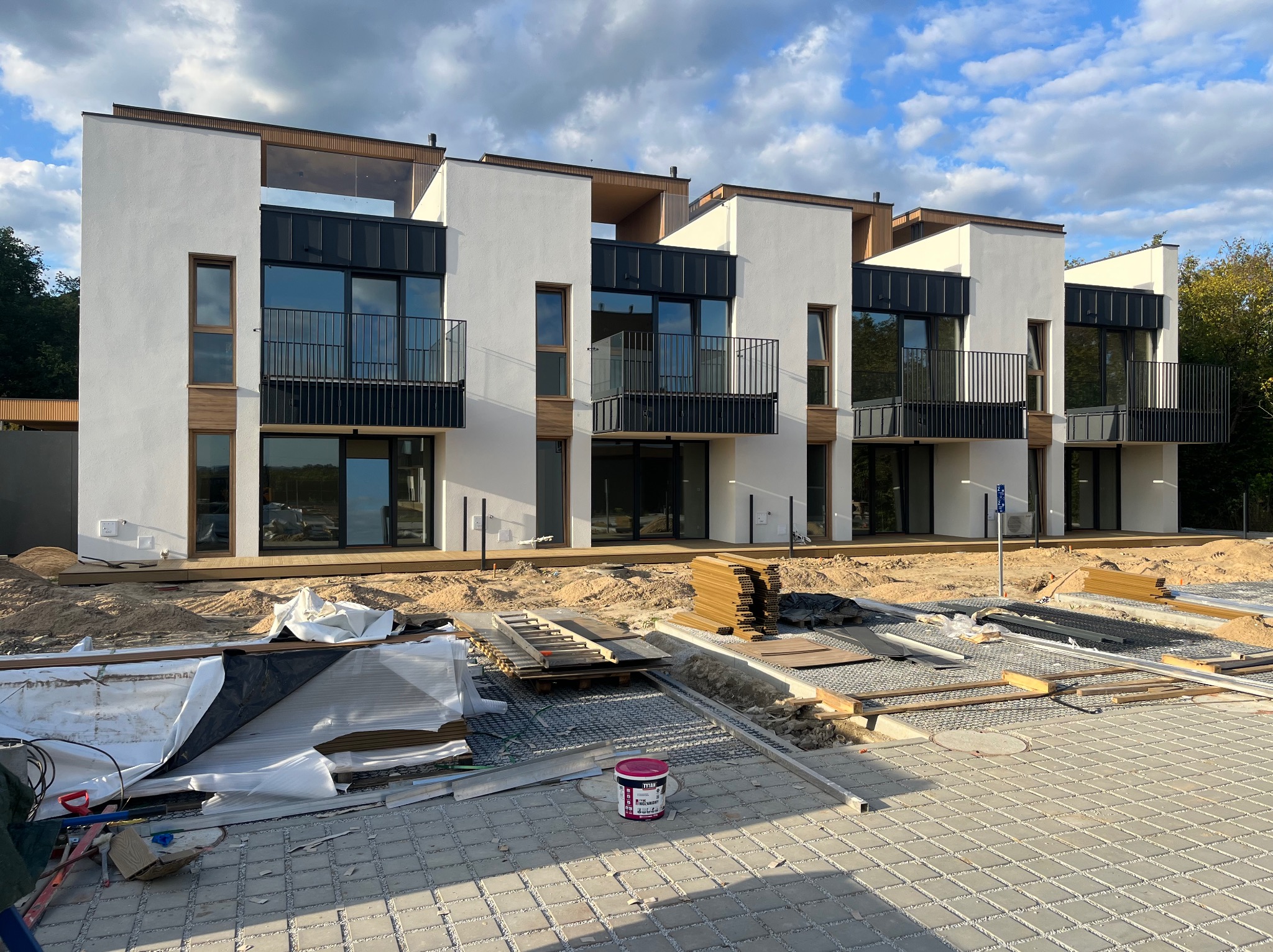 Montaż tarasu przy nowoczesnym domu szeregowym. Widoczne deski tarasowe, piasek i narzędzia. Etap budowy tarasu.