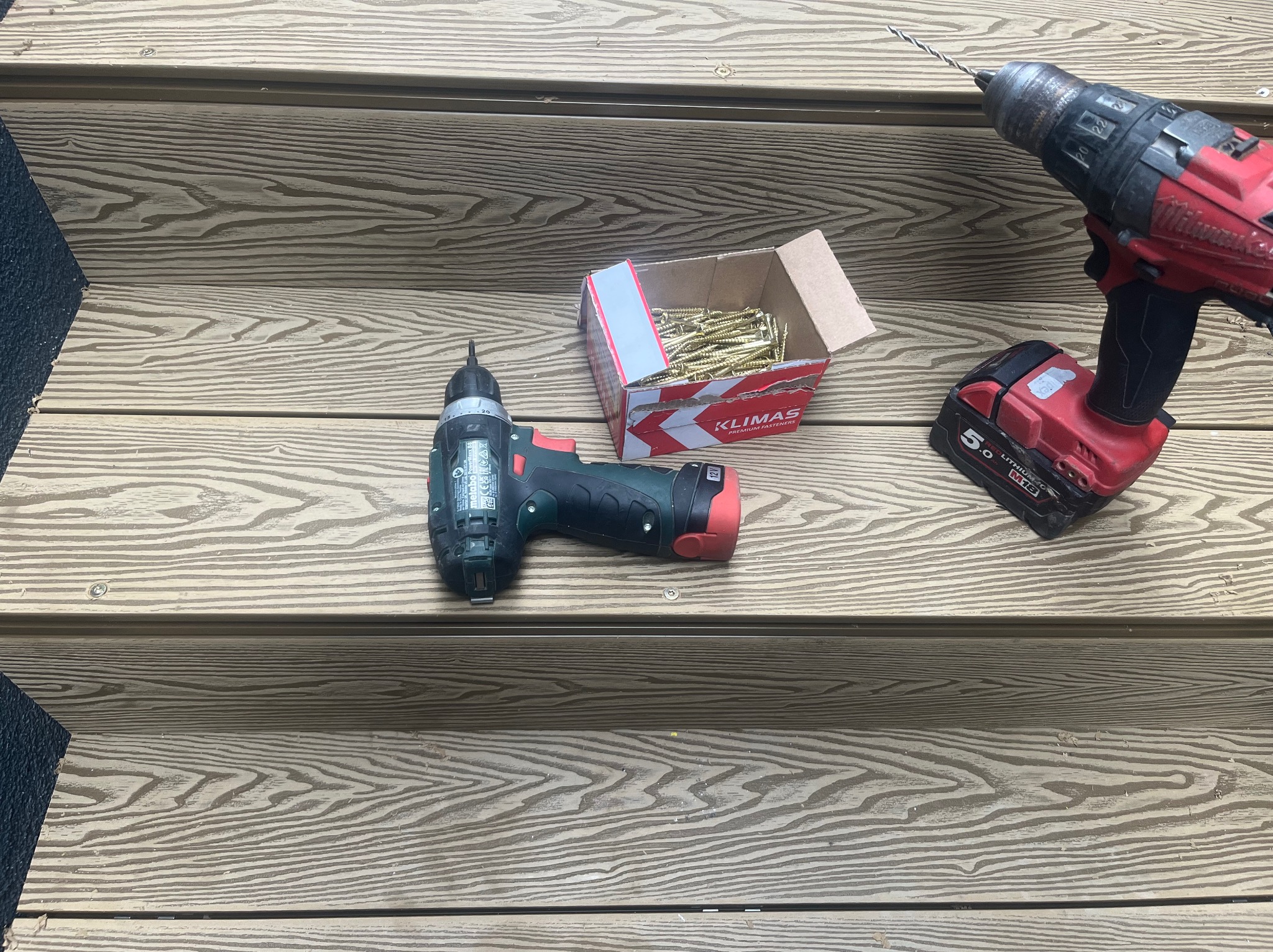 Montaż tarasu kompozytowego: wkrętarka Milwaukee z wiertłem, wkrętarka Metabo i otwarte pudełko wkrętów Klimas leżą na desce tarasowej o teksturze drewna.