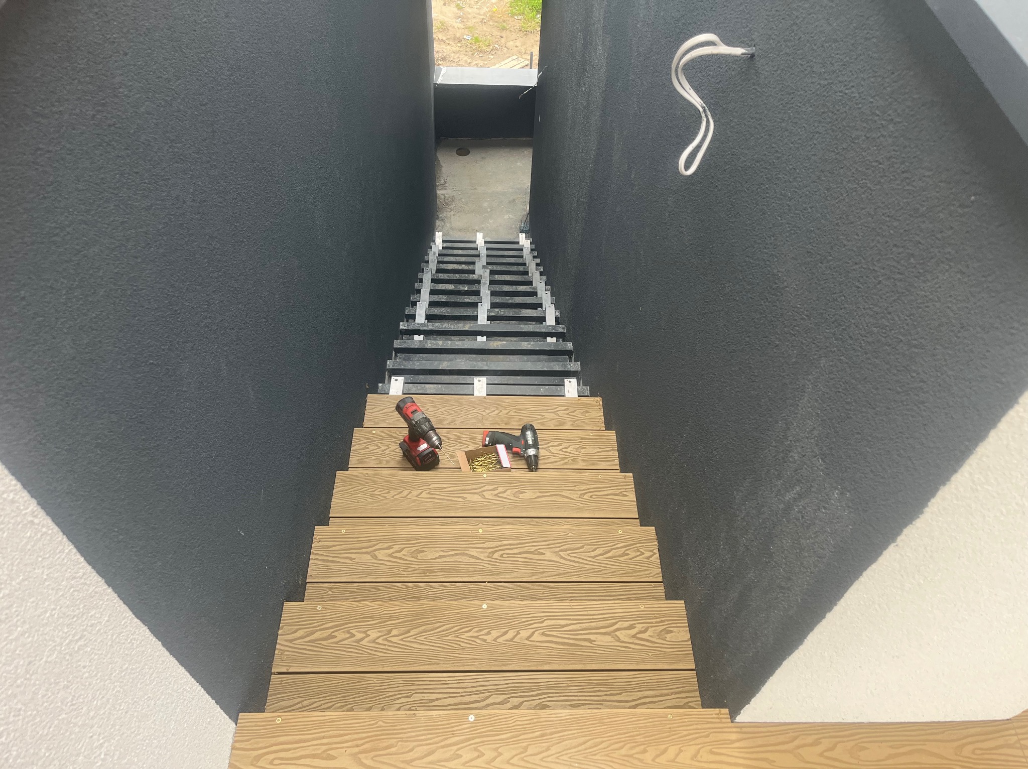 Montaż schodów z desek na metalowym stelażu. Widoczne wkrętarki i wkręty. Ciemne ściany tworzą tunel prowadzący na zewnątrz. Ujęcie z góry.