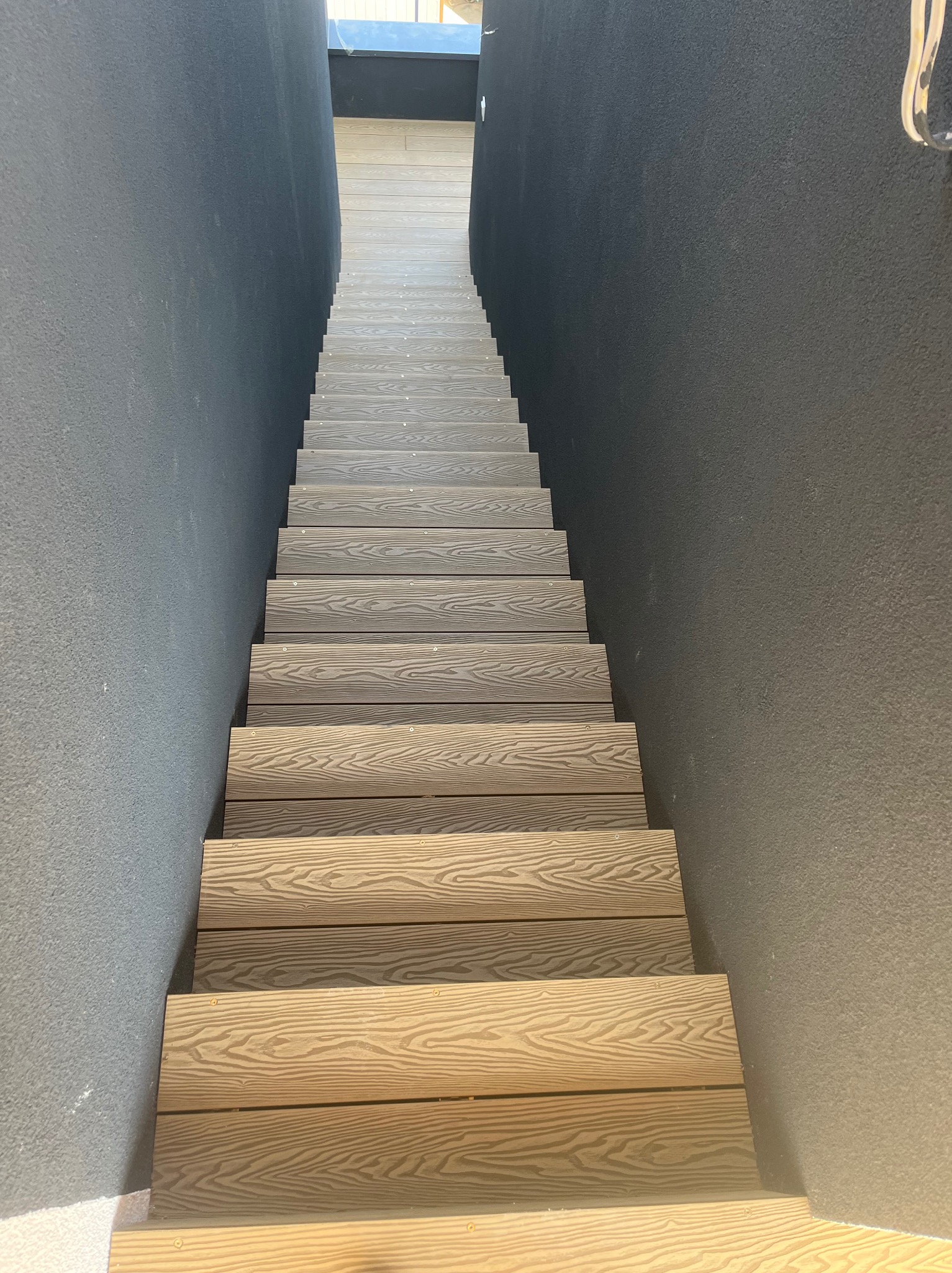Schody z desek kompozytowych w wąskim przejściu. Deski w odcieniach beżu i brązu, ułożone pomiędzy ścianami o szarej, chropowatej fakturze. Widok z góry.
