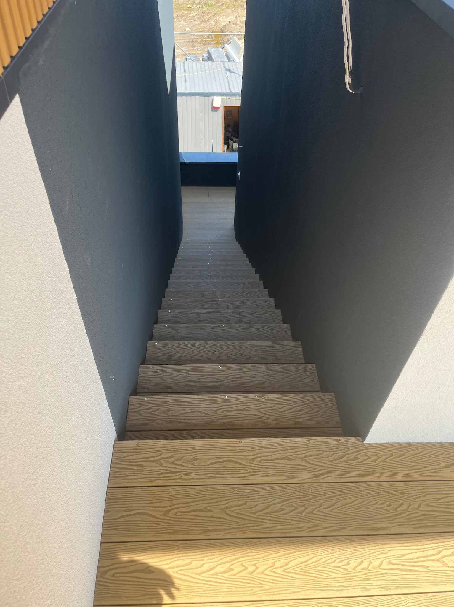 Schody tarasowe z desek o fakturze drewna, osadzone między białymi i grafitowymi ścianami. Widok z góry na stopnie.