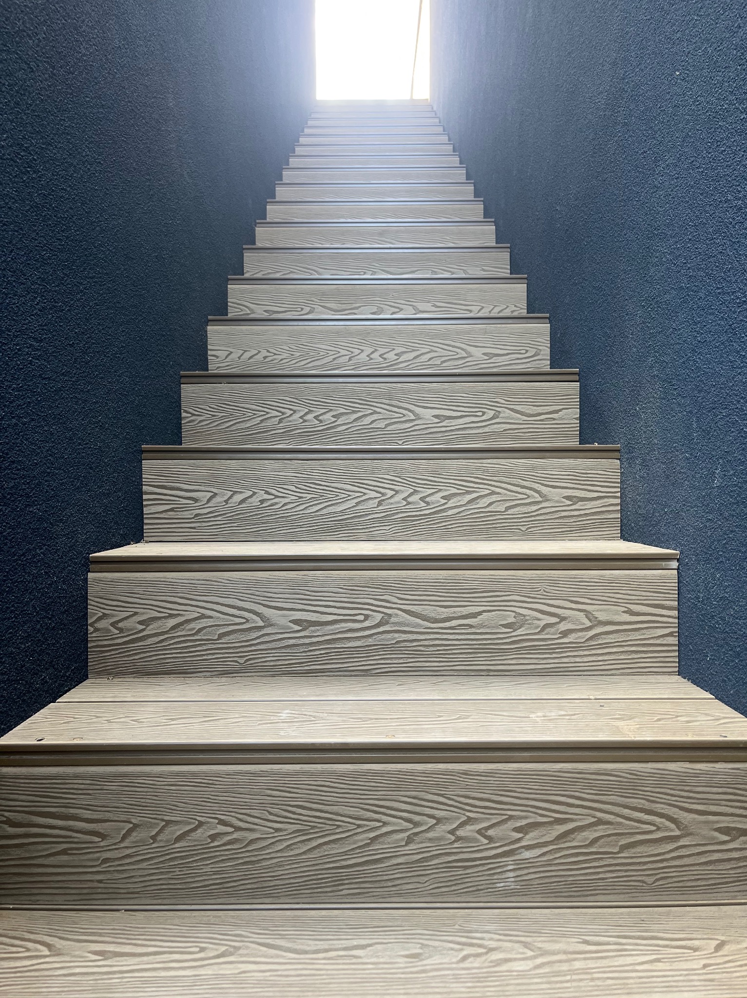 Schody zewnętrzne z desek o teksturze drewna, prowadzące ku górze, na tle ciemnej ściany. Widok z dołu.