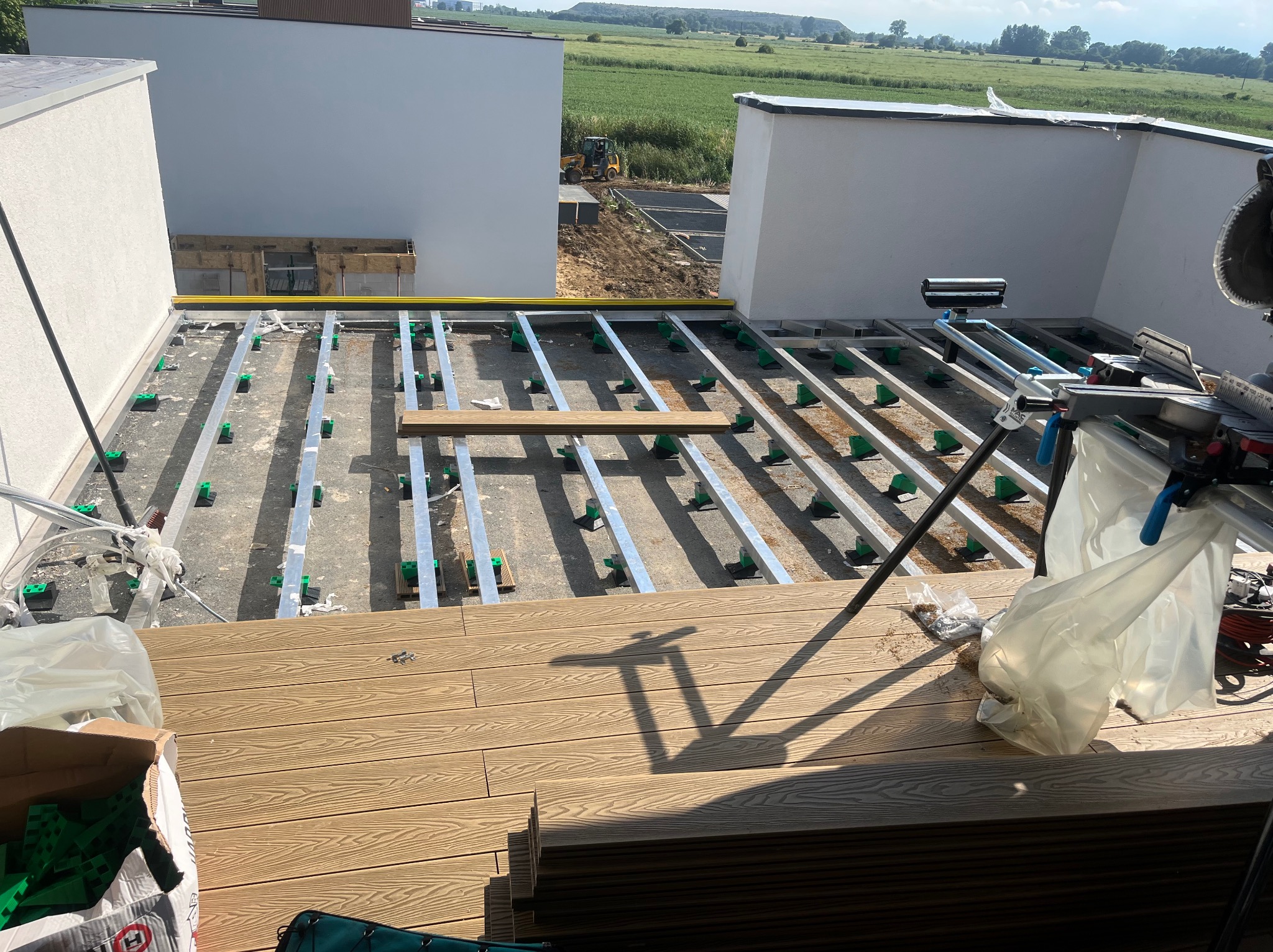 Montaż tarasu: widok na częściowo ułożoną konstrukcję z legarów i wsporników, w tle budynek i zieleń. U góry widoczne narzędzia pracy.