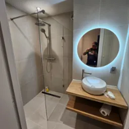 Nowoczesna łazienka z kabiną prysznicową typu walk-in, umywalką nablatową na drewnianym blacie z podświetlanym lustrem i beżowymi płytkami na ścianach i podłodze.