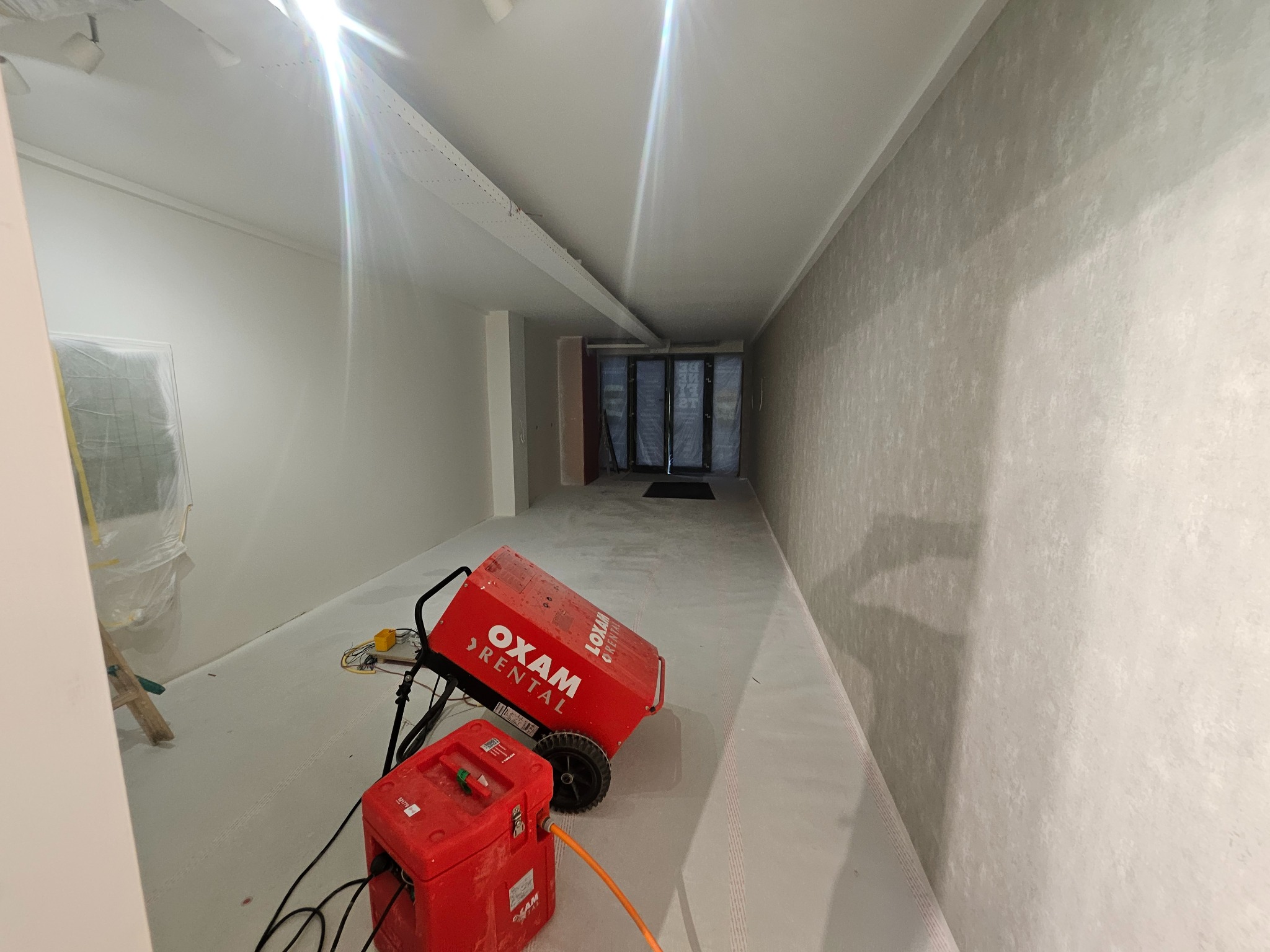 Wnętrze w trakcie remontu: świeżo pomalowane ściany w odcieniach bieli i szarości, z widocznym nagrzewnicą OXAM Rental i drabina zabezpieczona folią malarską.