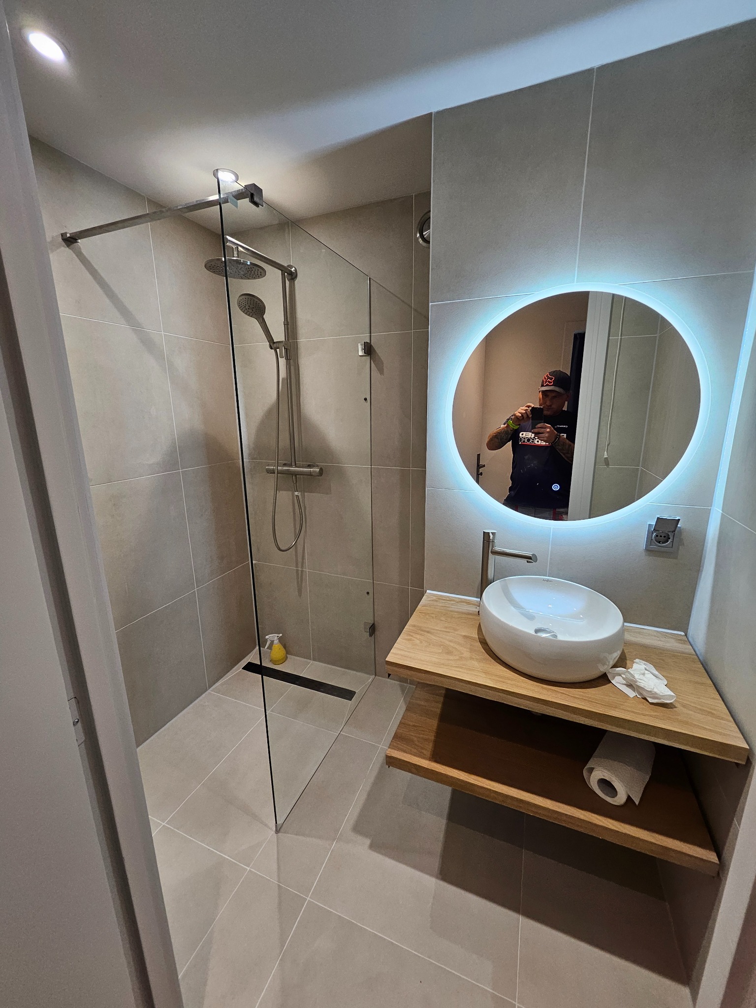 Nowoczesna łazienka z kabiną prysznicową typu walk-in, umywalką nablatową na drewnianym blacie z podświetlanym lustrem i beżowymi płytkami na ścianach i podłodze.