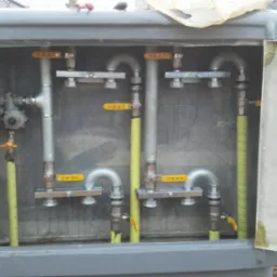 Szara szafka z otwartymi drzwiczkami, wewnątrz instalacja gazowa z rurami, zaworami z żółtymi uchwytami i reduktorem, na zewnątrz widoczny fragment trawnika i ogrodzenia z siatki.