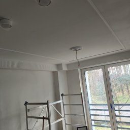 USŁUGI I HANDEL DAMIAN ZDRENKA - Wnętrze w trakcie remontu: widok na sufit z dekoracyjnymi listwami, zamontowanymi lampami, wiszącymi kablami i rozstawionym rusztowaniem przy oknie.