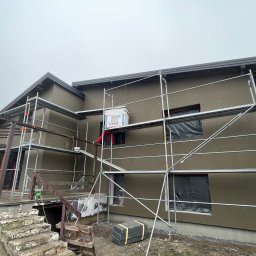 USŁUGI I HANDEL DAMIAN ZDRENKA - Dom jednorodzinny w trakcie ocieplania, widoczne rusztowania, okna zabezpieczone folią, paczka styropianu na rusztowaniu, elewacja w kolorze beżowym.