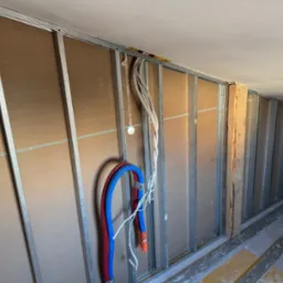 Konstrukcja ścian działowych z profili stalowych i płyt, widoczne przewody instalacji elektrycznej i rury instalacji wodnej, fragment podłogi z żółtymi plamami.