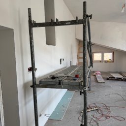 USŁUGI I HANDEL DAMIAN ZDRENKA - Wnętrze w trakcie prac wykończeniowych: metalowe rusztowanie, ściany po szpachlowaniu, widoczne puszki elektryczne, podłoga z resztkami materiałów budowlanych, okno dachowe w tle.