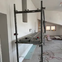 Wnętrze w trakcie prac wykończeniowych: metalowe rusztowanie, ściany po szpachlowaniu, widoczne puszki elektryczne, podłoga z resztkami materiałów budowlanych, okno dachowe w tle.