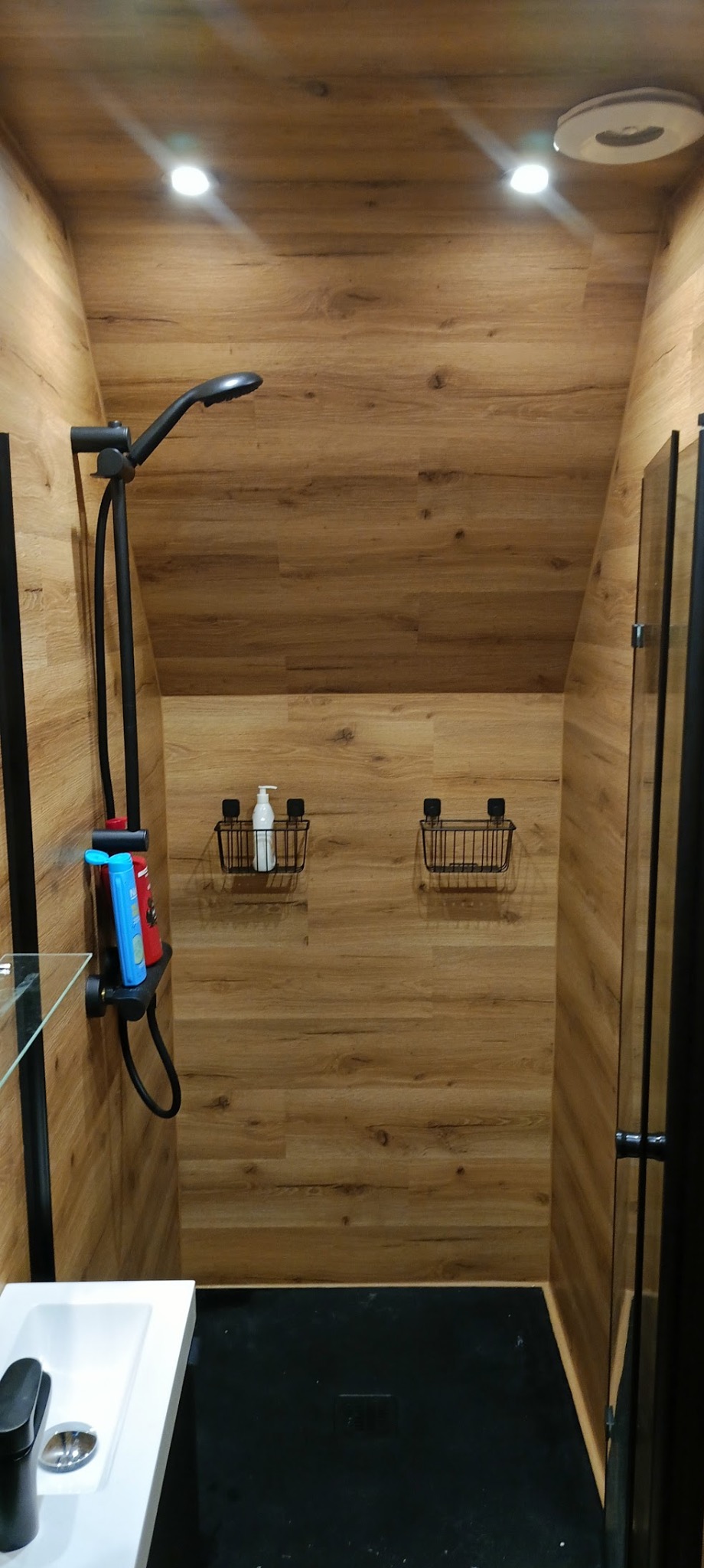 Nowoczesna kabina prysznicowa z drewnopodobnymi panelami na ścianach, czarną armaturą i półkami z czarnego drutu, oświetlona punktowymi lampami sufitowymi.