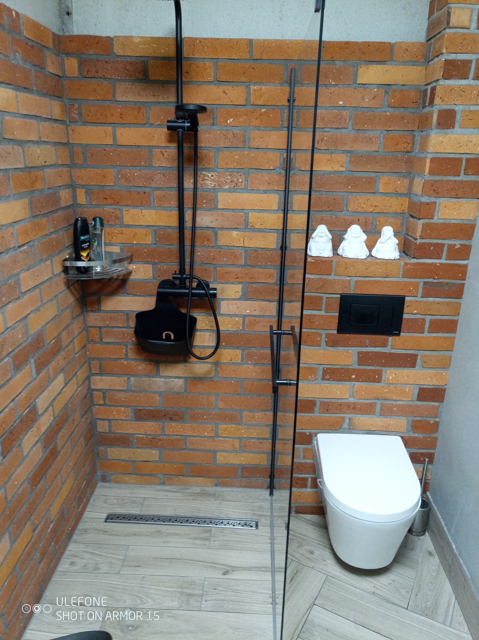 Nowoczesna łazienka z kabiną prysznicową typu walk-in, wykończona cegłą, z czarną armaturą i drewnopodobną podłogą, minimalistyczne figurki na ścianie.