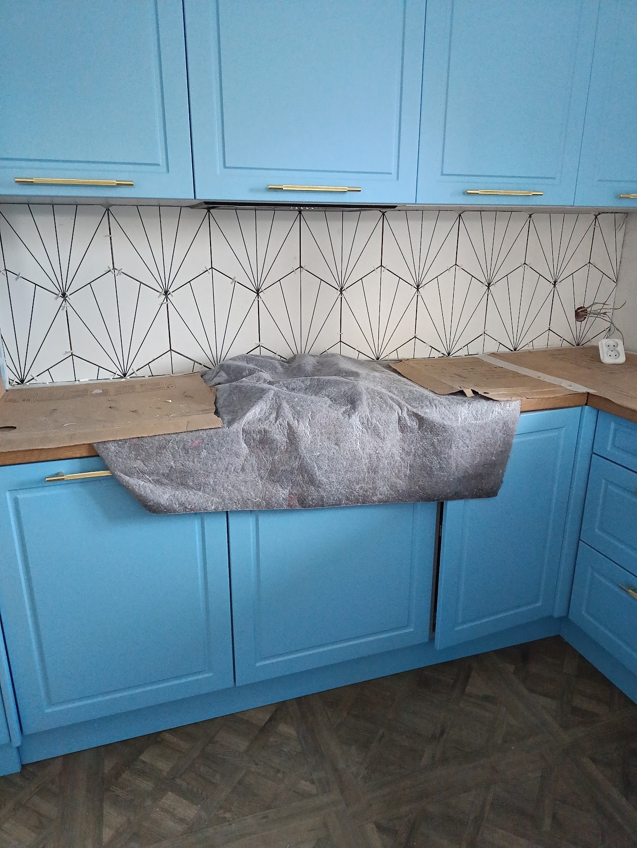 Niebieska zabudowa kuchenna z złotymi uchwytami, zabezpieczony blat i geometryczne płytki na ścianie w trakcie montażu, widoczne gniazdko elektryczne i kable.