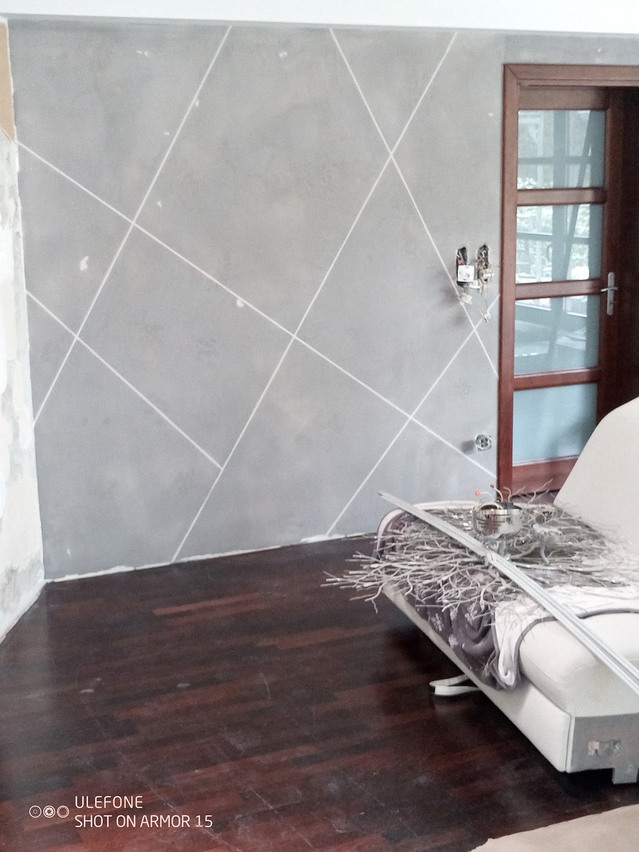 Szara ściana z geometrycznym wzorem z białych linii, widoczne niedokończone wykończenia przy podłodze i gniazdko elektryczne bez osłony, drewniana podłoga i fragment białej kanapy z dekoracją.