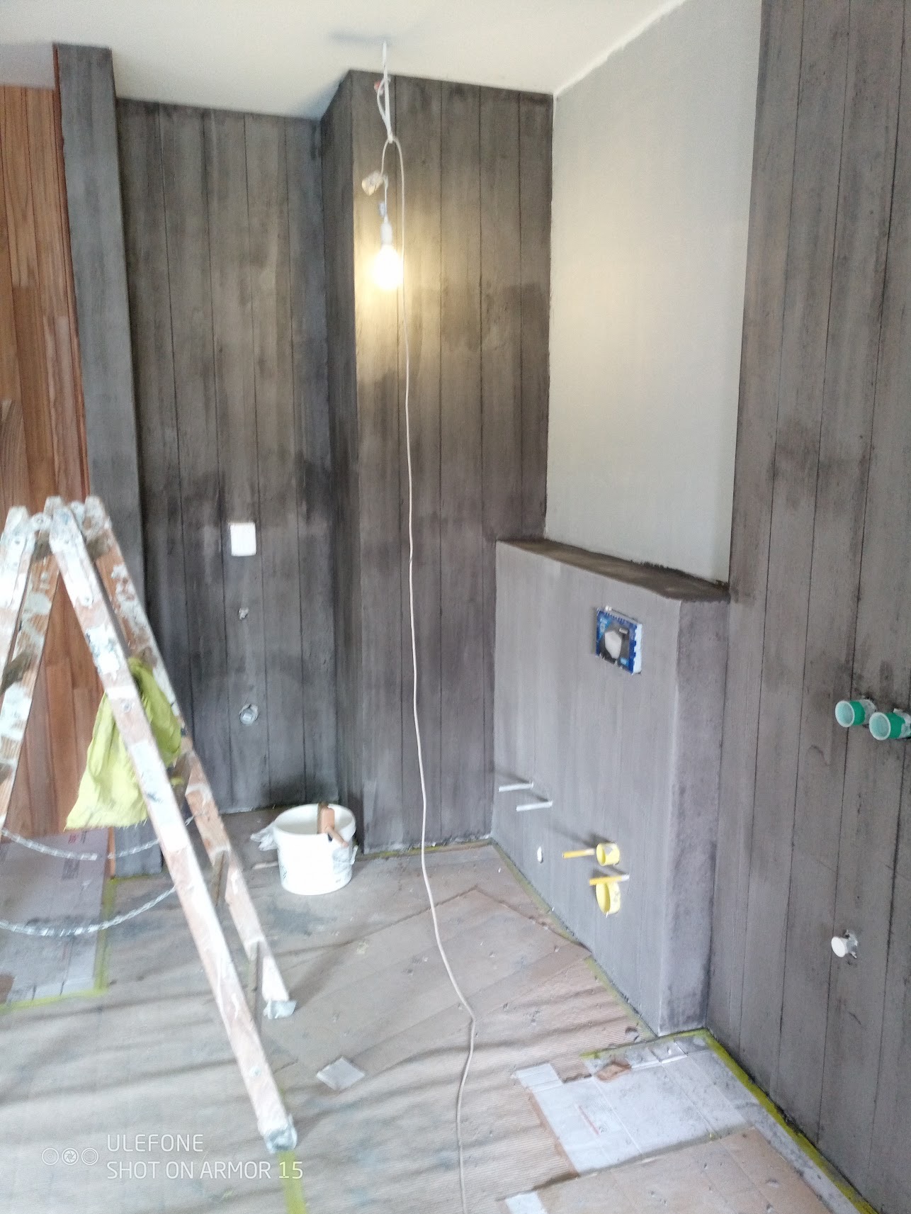 Łazienka w trakcie remontu: ściany wykończone panelami imitującymi beton, widoczne instalacje wodno-kanalizacyjne, drabina malarska z plamami farby i wiadro z narzędziami.