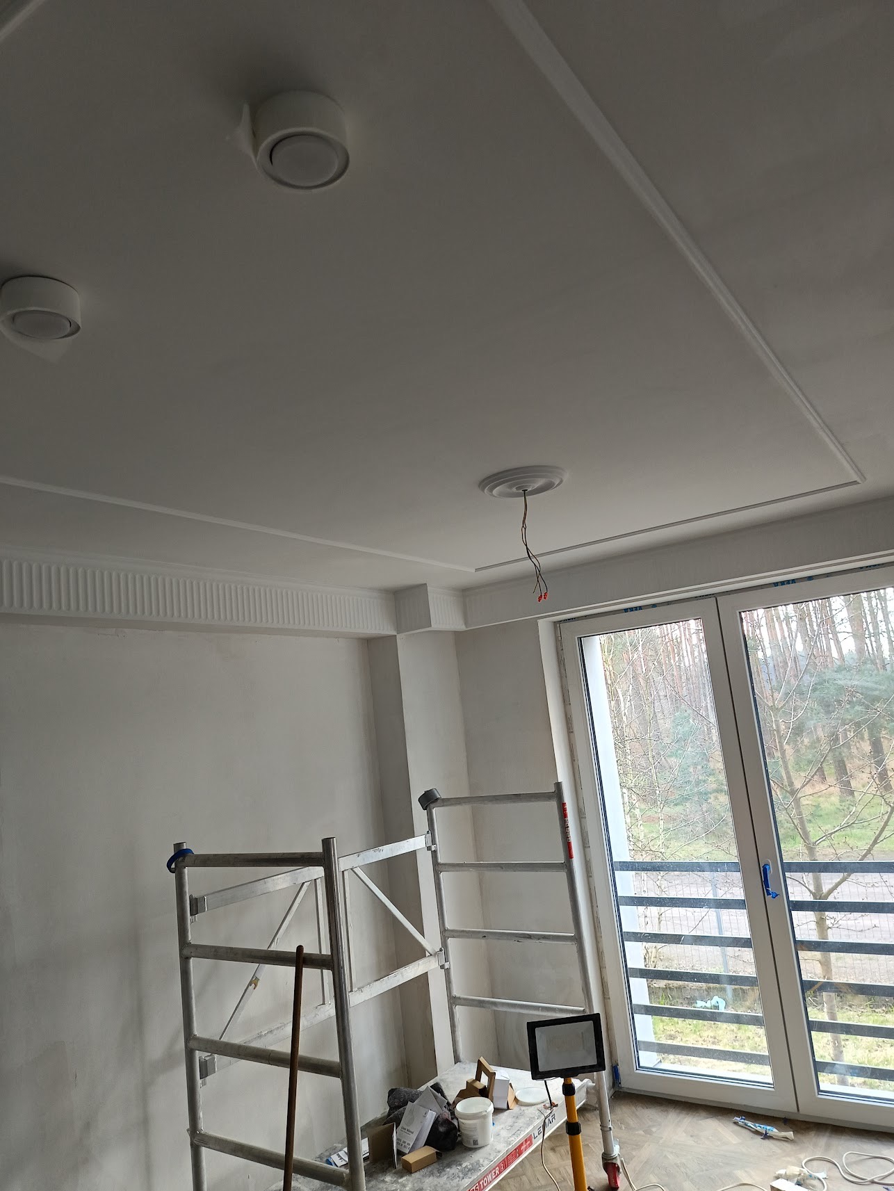 Wnętrze w trakcie remontu: widok na sufit z dekoracyjnymi listwami, zamontowanymi lampami, wiszącymi kablami i rozstawionym rusztowaniem przy oknie.